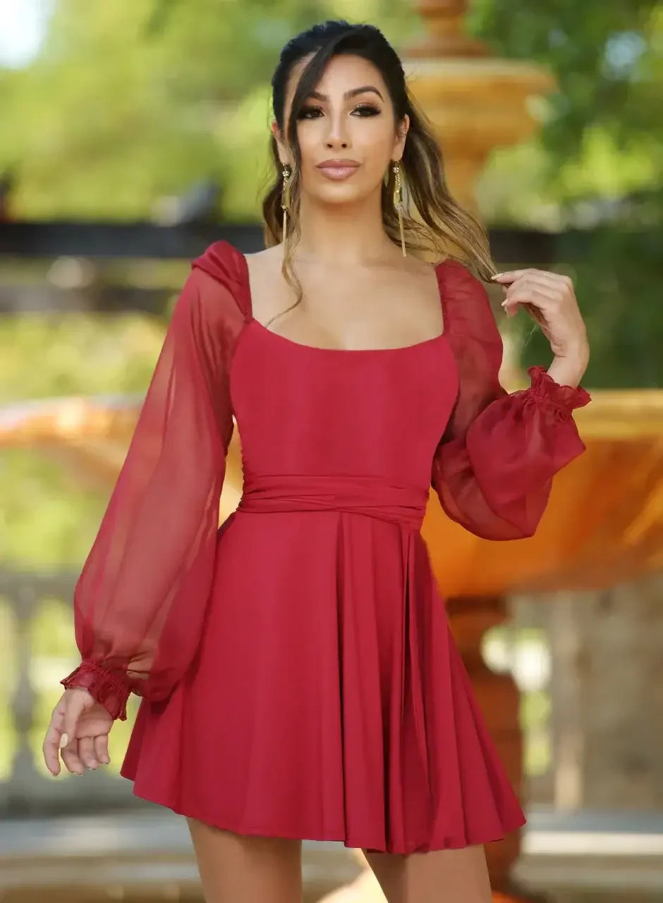 A woman poses confidently in a vibrant red dress with sheer, puffed sleeves, gently holding her hair while standing against a blurred background of greenery and a fountain.