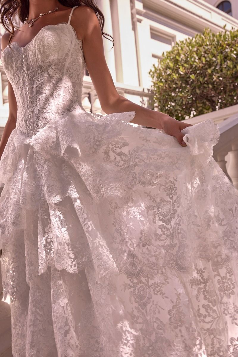 WL076 - LAYERED SCALLOP LACE A-LINE GOWN - Andrea & Leo | Lady Black Tie | A close-up of a woman wearing a white lace wedding gown, holding out the skirt to show its fullness. The dress features a fitted lace bodice, thin straps, and a layered ruffled skirt.