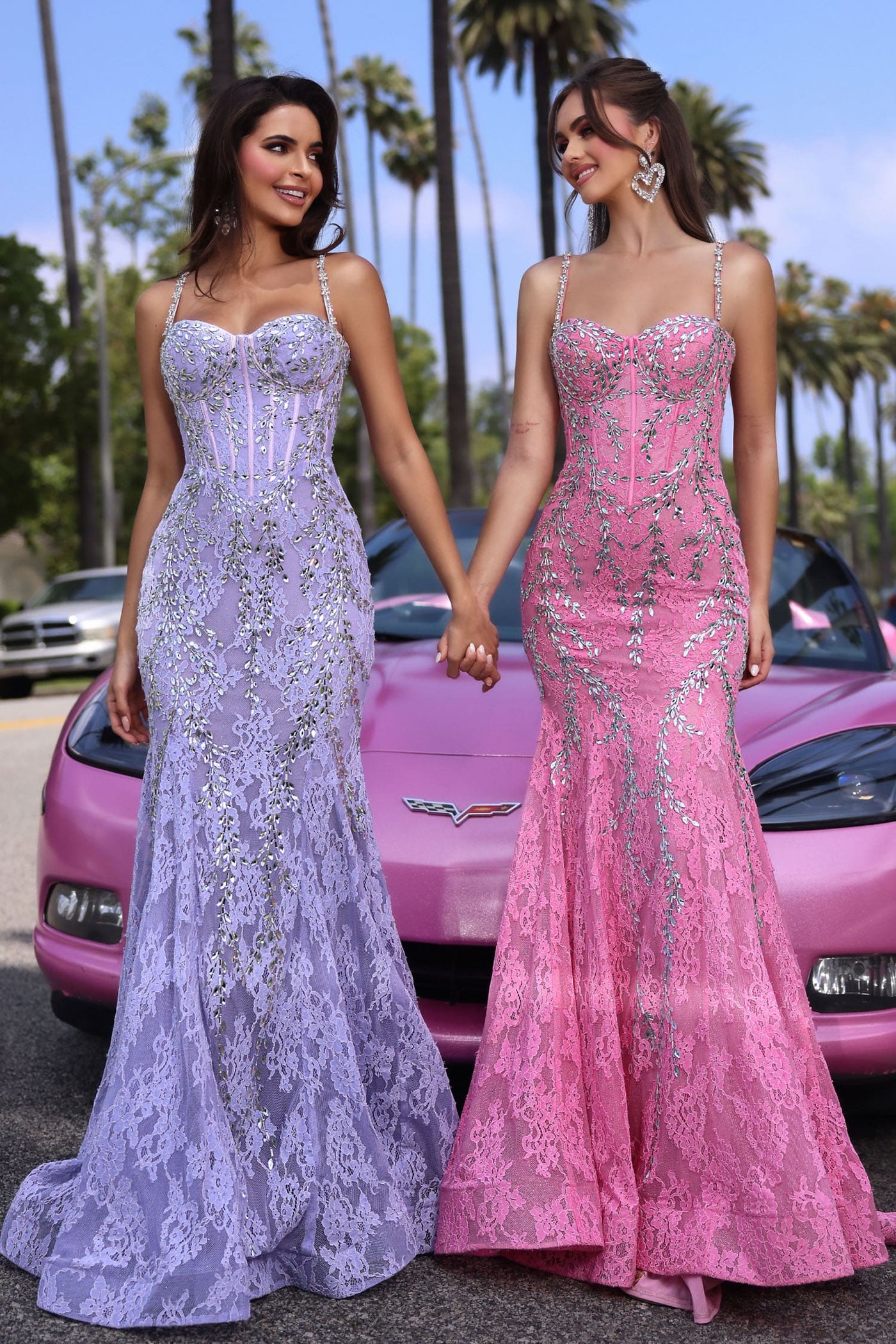 Two women in elegant evening gowns standing in front of a pink car with palm trees in the background.