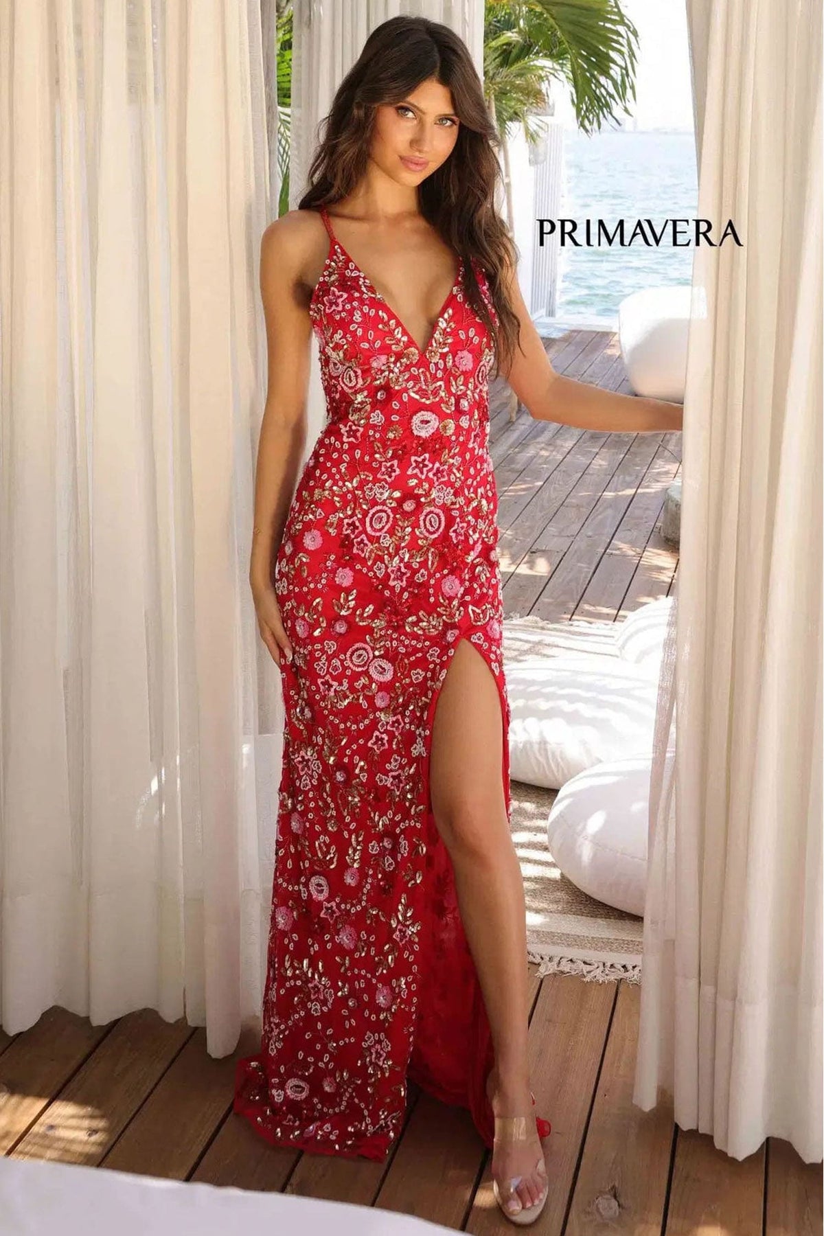 Woman wearing a red floral dress standing by a wooden deck with curtains and a view of the ocean.