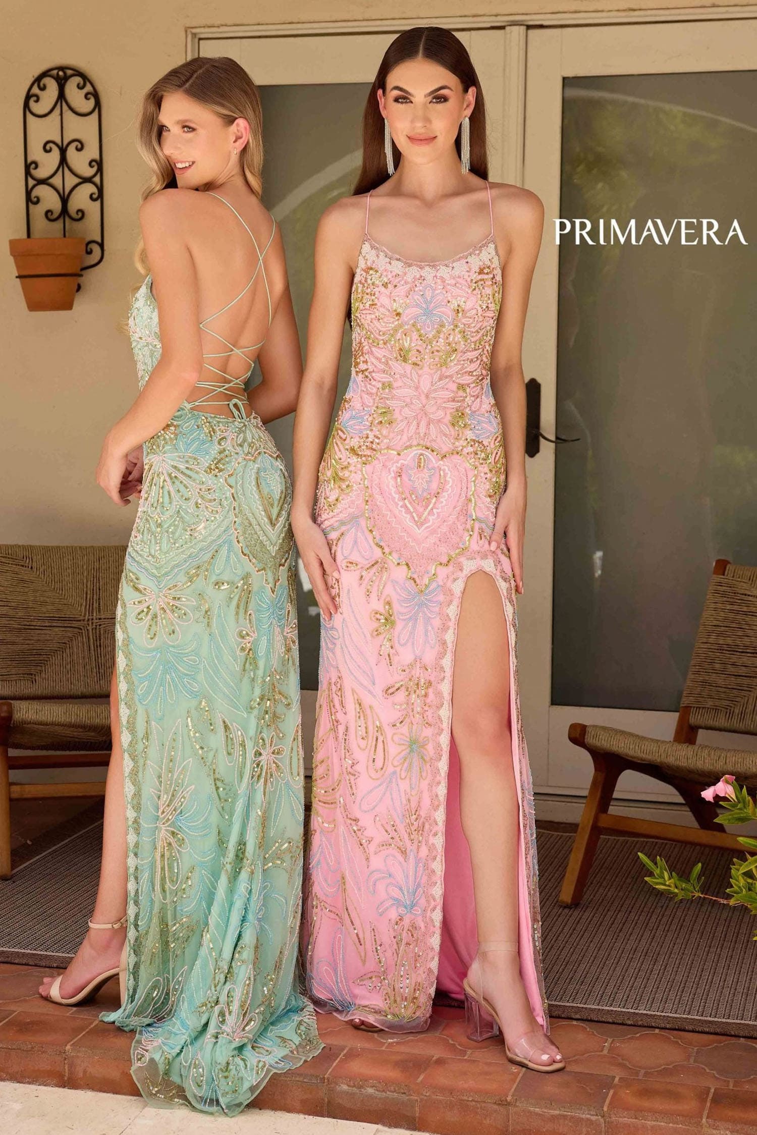 Two models wearing elegant evening gowns in an outdoor patio setting. The model on the left wears a light aqua green gown with gold embroidery and an open back, while the model on the right wears a pink gown with gold embroidery and a thigh-high slit.