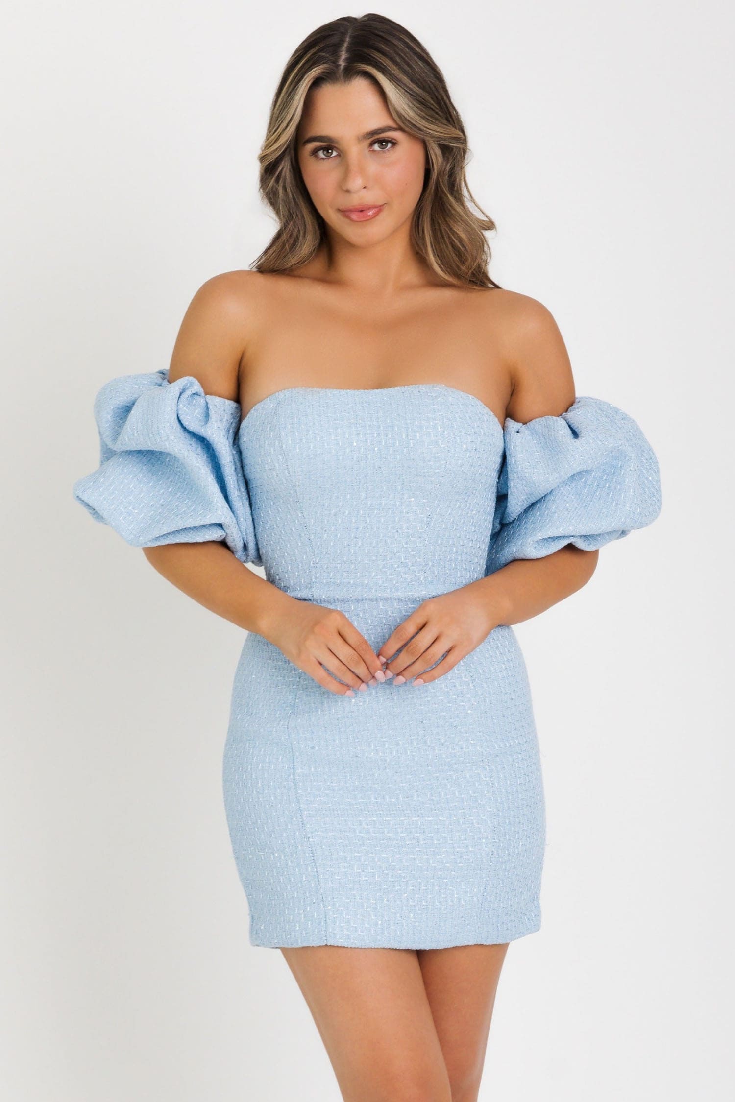 A woman modeling a light blue off-the-shoulder mini dress with textured fabric and voluminous puff sleeves, photographed against a plain light gray studio background.