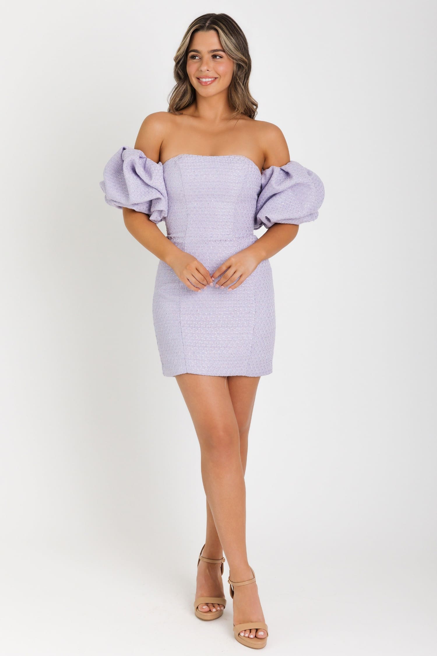 A woman modeling a lavender off-the-shoulder mini dress with voluminous puff sleeves and a subtle textured pattern, paired with beige high-heeled sandals against a plain white studio background.