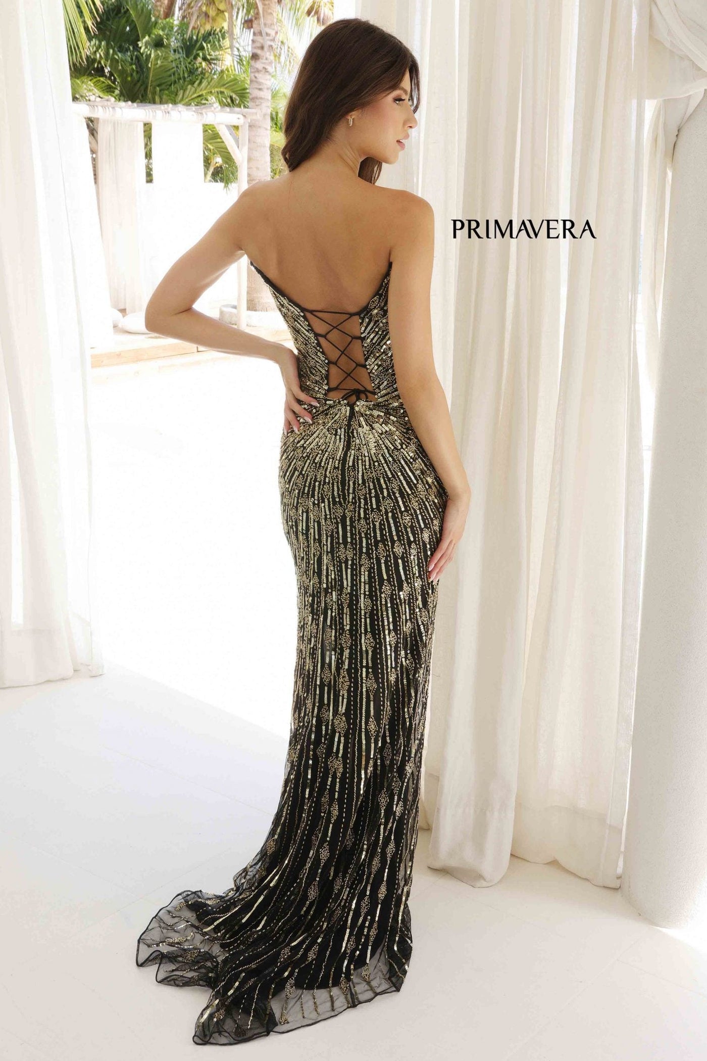 Woman wearing a black evening gown with intricate detailing, standing in front of white curtains.