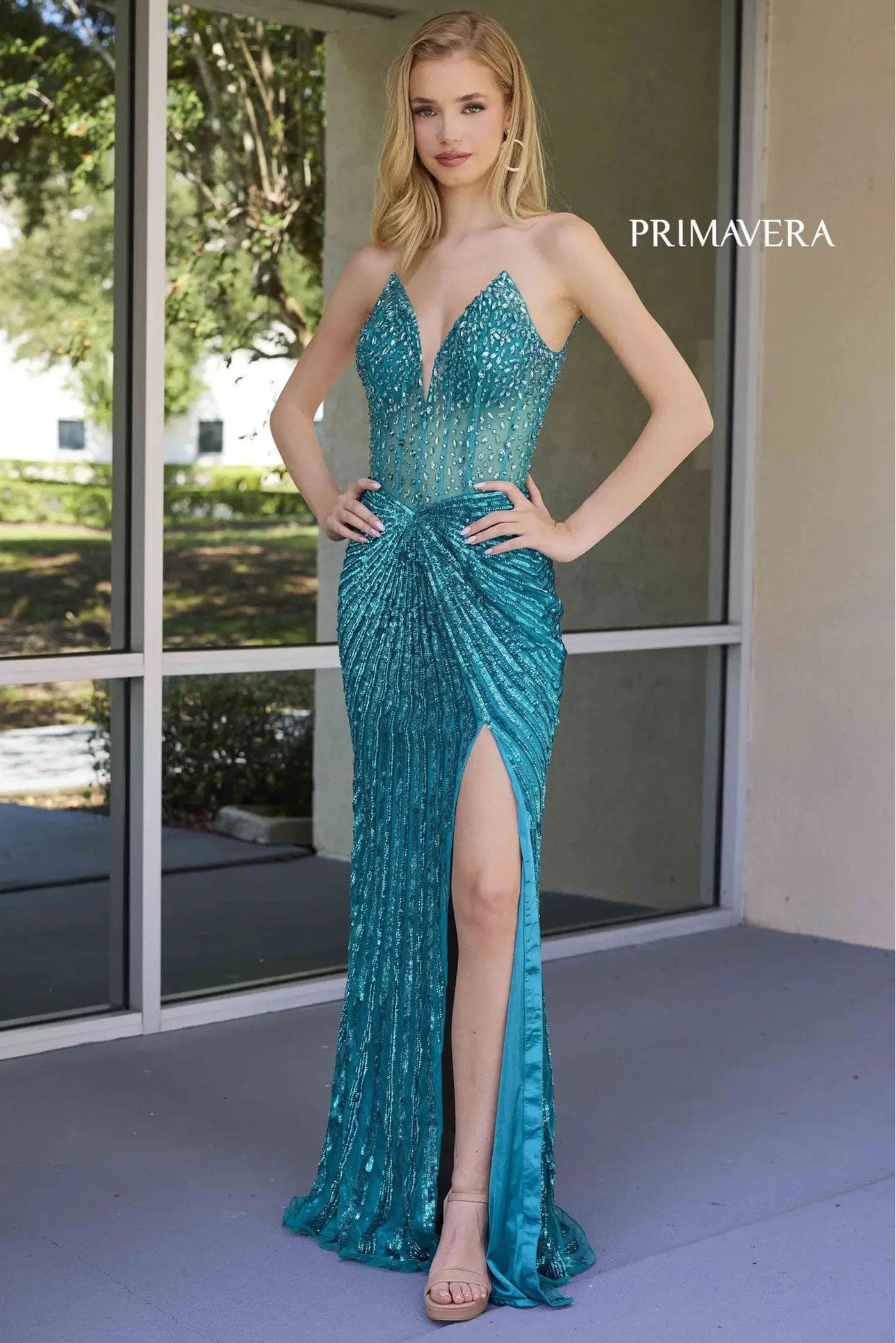 Woman wearing a teal evening gown with a high slit, standing in front of a glass door.