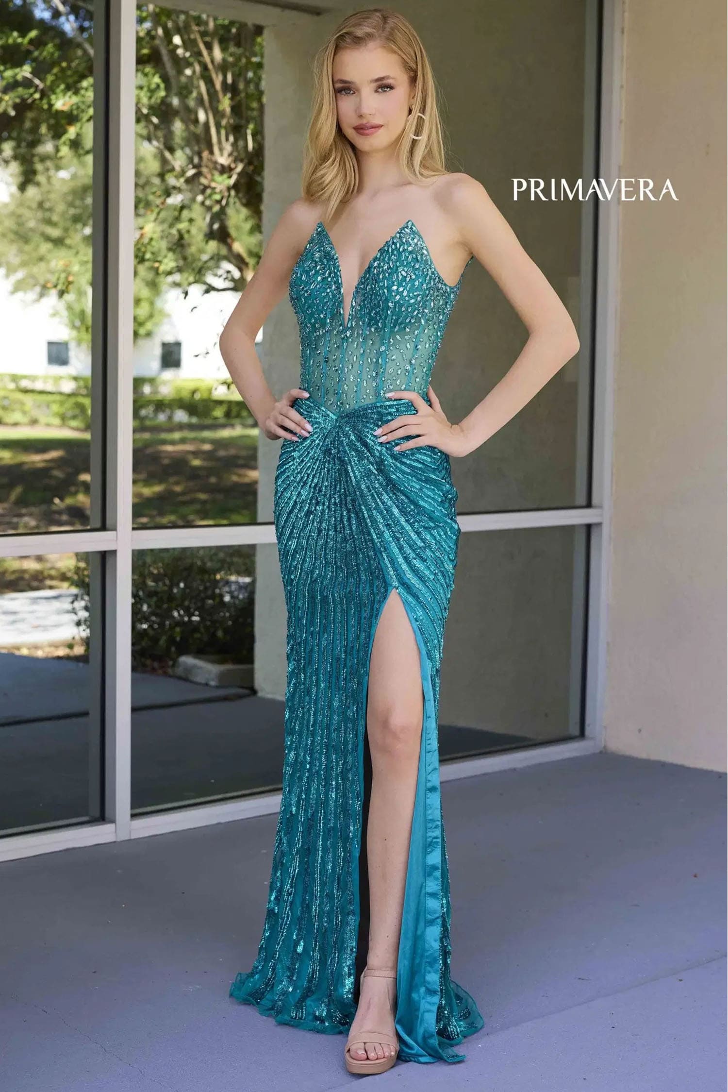 Woman wearing a teal evening gown with a high slit, standing in front of a glass door.