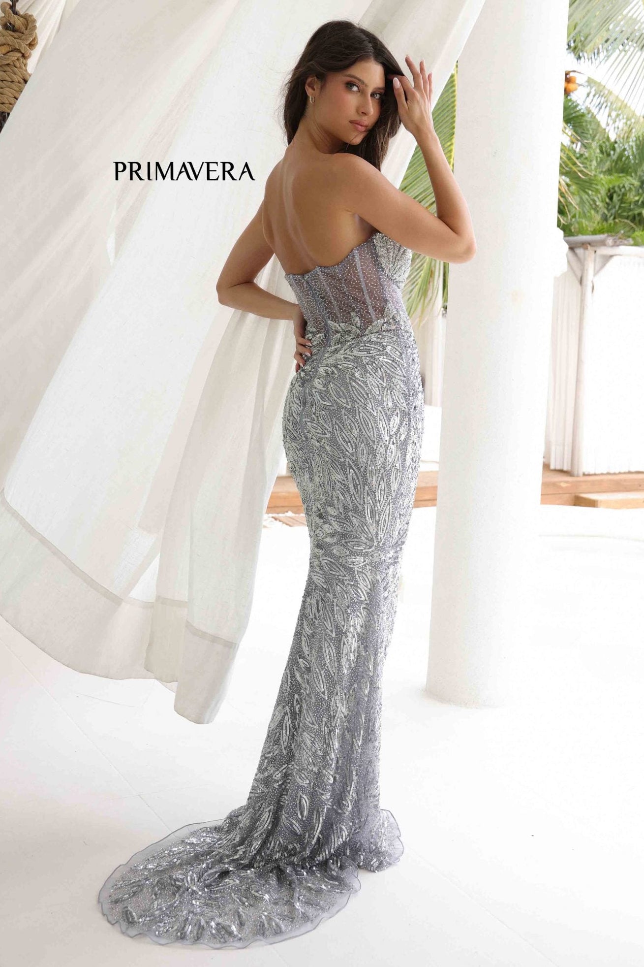 Woman in a silver evening gown with 'Primavera' branding, standing in a tropical setting.