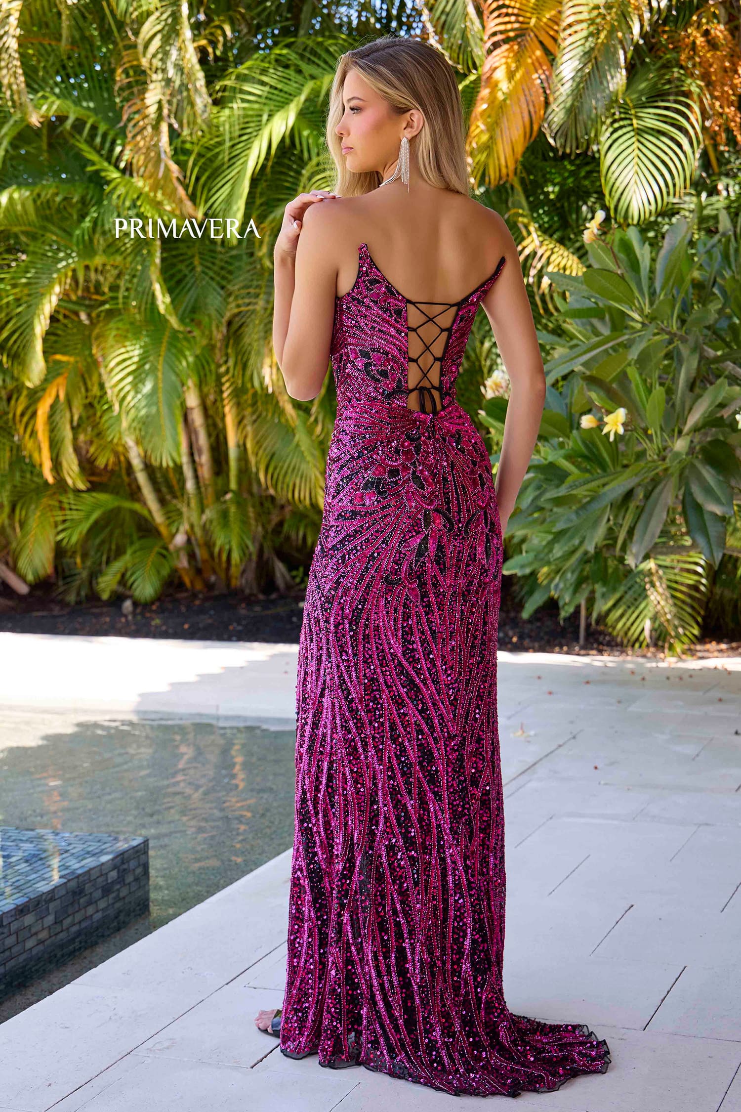 A woman stands outdoors by a pool wearing a long magenta sequined evening gown with thin straps and an open lace-up back. Palm foliage and a patio are visible in the background, and the brand name Primavera appears on the left side.
