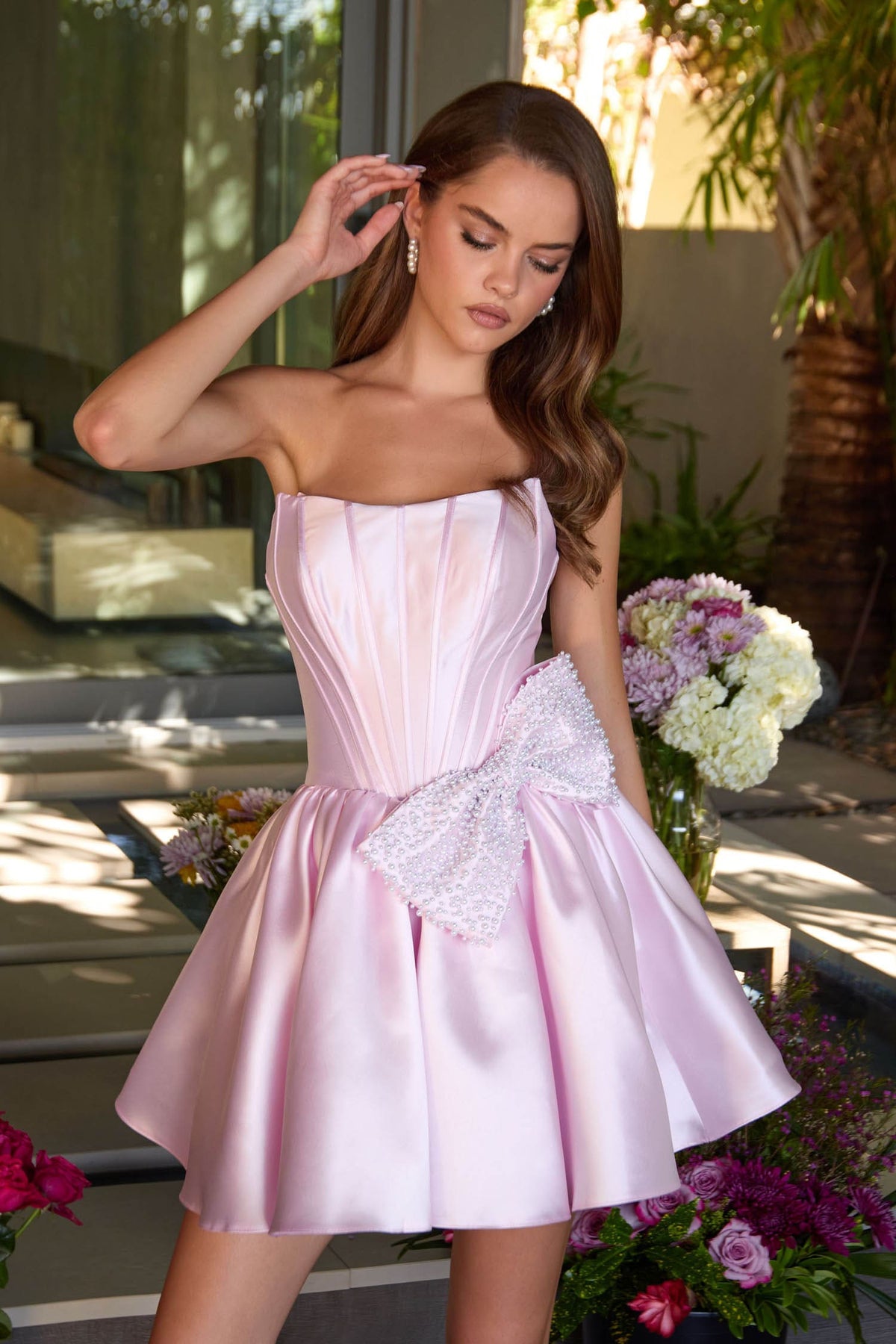 Woman in a pink strapless dress with a bow, standing outdoors with flowers and a building in the background.