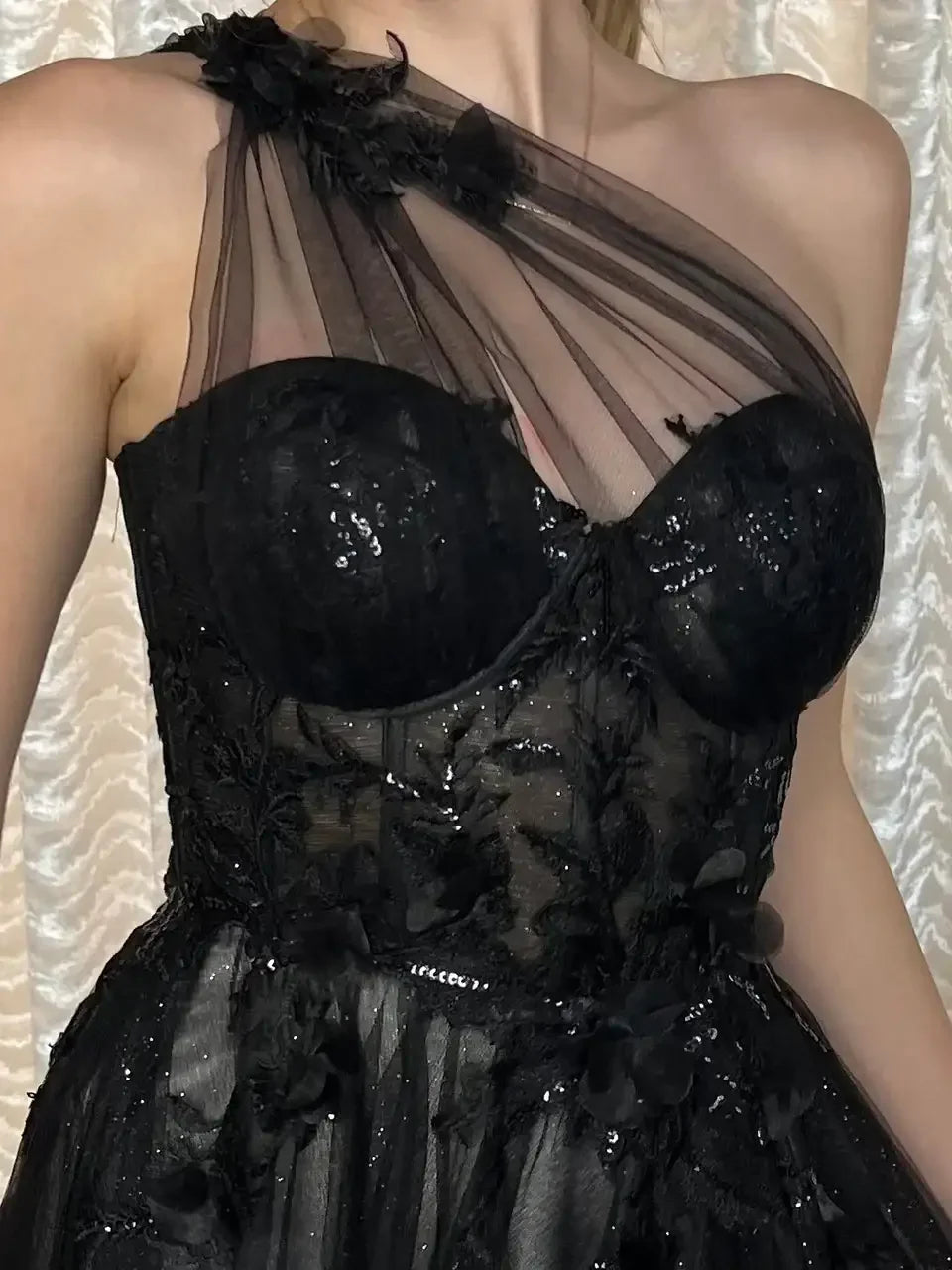 A black evening dress features a fitted bodice with intricate lace and sequin detailing. The sheer overlay drapes elegantly from one shoulder, set against a softly lit, textured background.