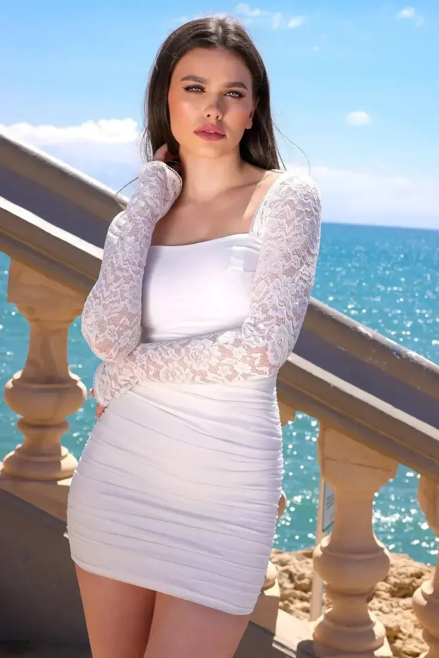 A woman in a white dress with lace sleeves stands by a stair railing, looking at the camera. The background features a bright blue ocean and clear sky, conveying a serene, sunny day.