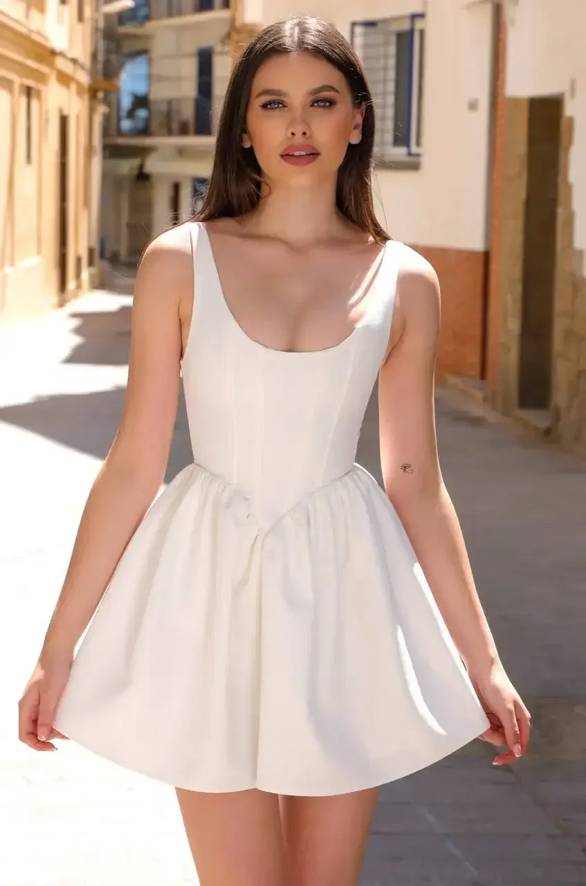 A young woman stands confidently, wearing a fitted white dress with a flared skirt. Sunlight illuminates a quaint street lined with buildings, enhancing the cheerful atmosphere.