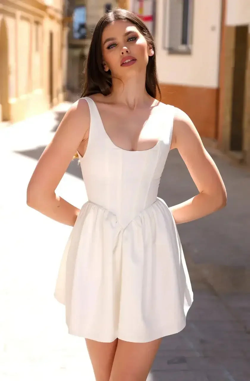 A young woman poses confidently in a fitted white dress with a flared skirt. Bright sunlight filters through a narrow, cobblestone street lined with warm-toned buildings.