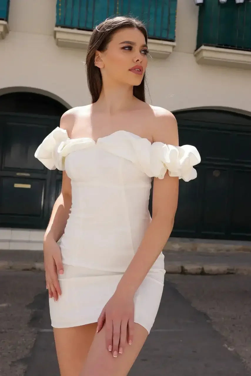 A woman in a fitted white dress with puffed sleeves stands confidently on a street. She gazes thoughtfully to her right, with a backdrop of green doors and light-colored buildings.