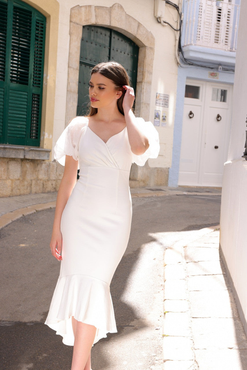 A woman in a white dress with puffed sleeves walks gracefully along a sunlit, narrow street, surrounded by stone buildings and green shutters, showcasing elegance in an outdoor setting.