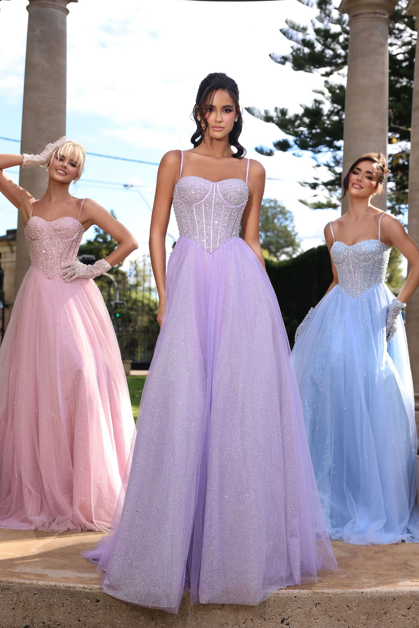 Three women modeling formal ball gowns outdoors under stone columns. The center model wears a lavender tulle gown with a beaded corset bodice, flanked by pink and light blue gowns.