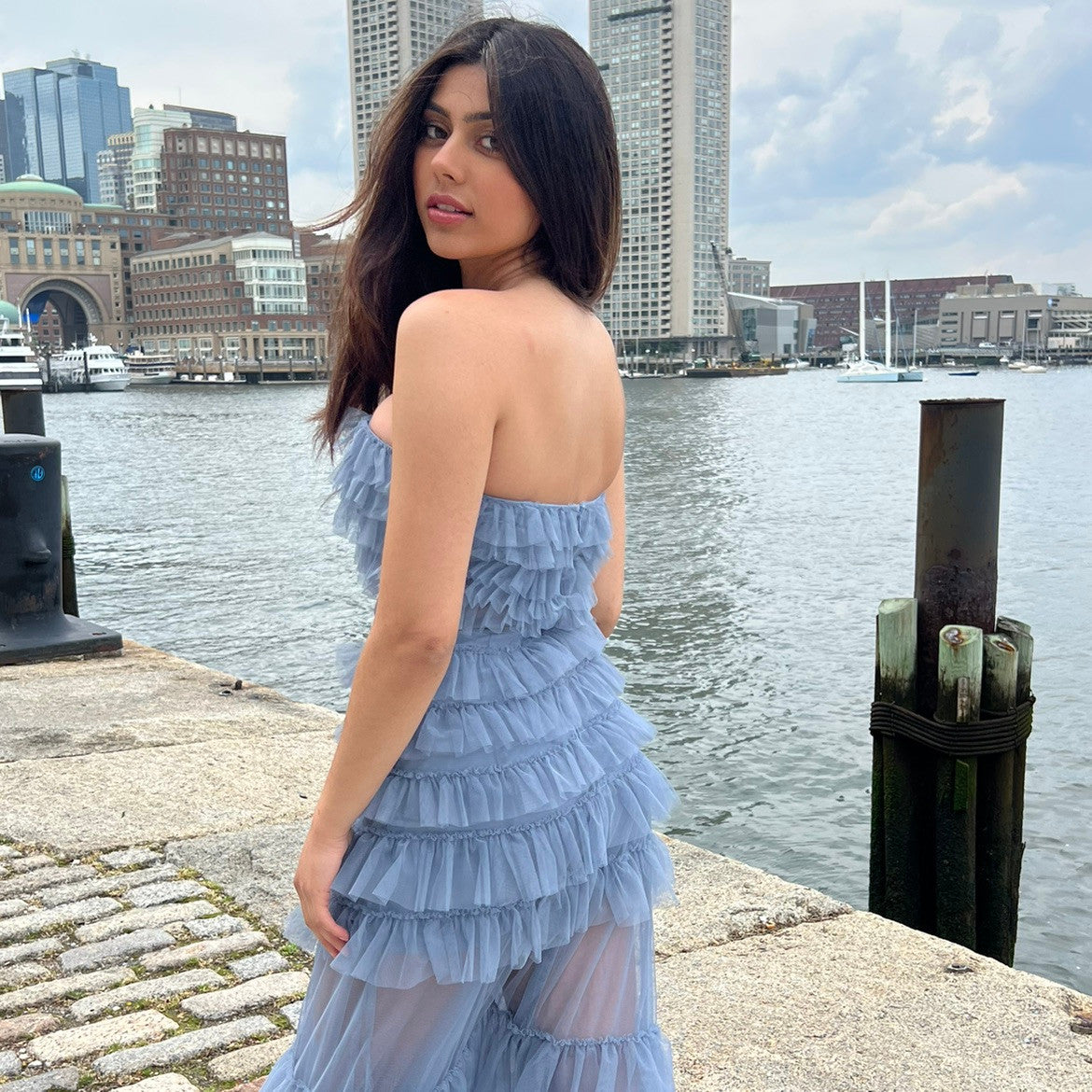 A woman in a layered, light blue dress stands by the water, looking back over her shoulder. The backdrop features tall buildings and boats, under a cloudy sky.