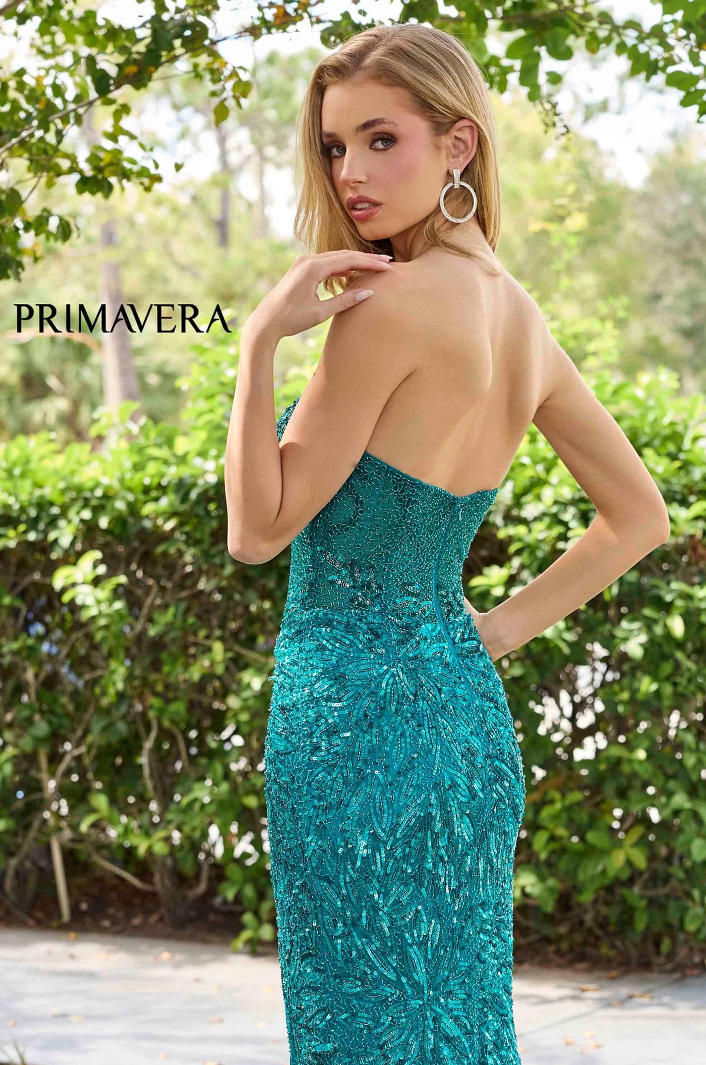 A woman wearing a strapless, fitted teal evening gown with a textured sequin or lace-like surface, photographed outdoors in front of green foliage. The brand name PRIMAVERA appears in the upper left.
