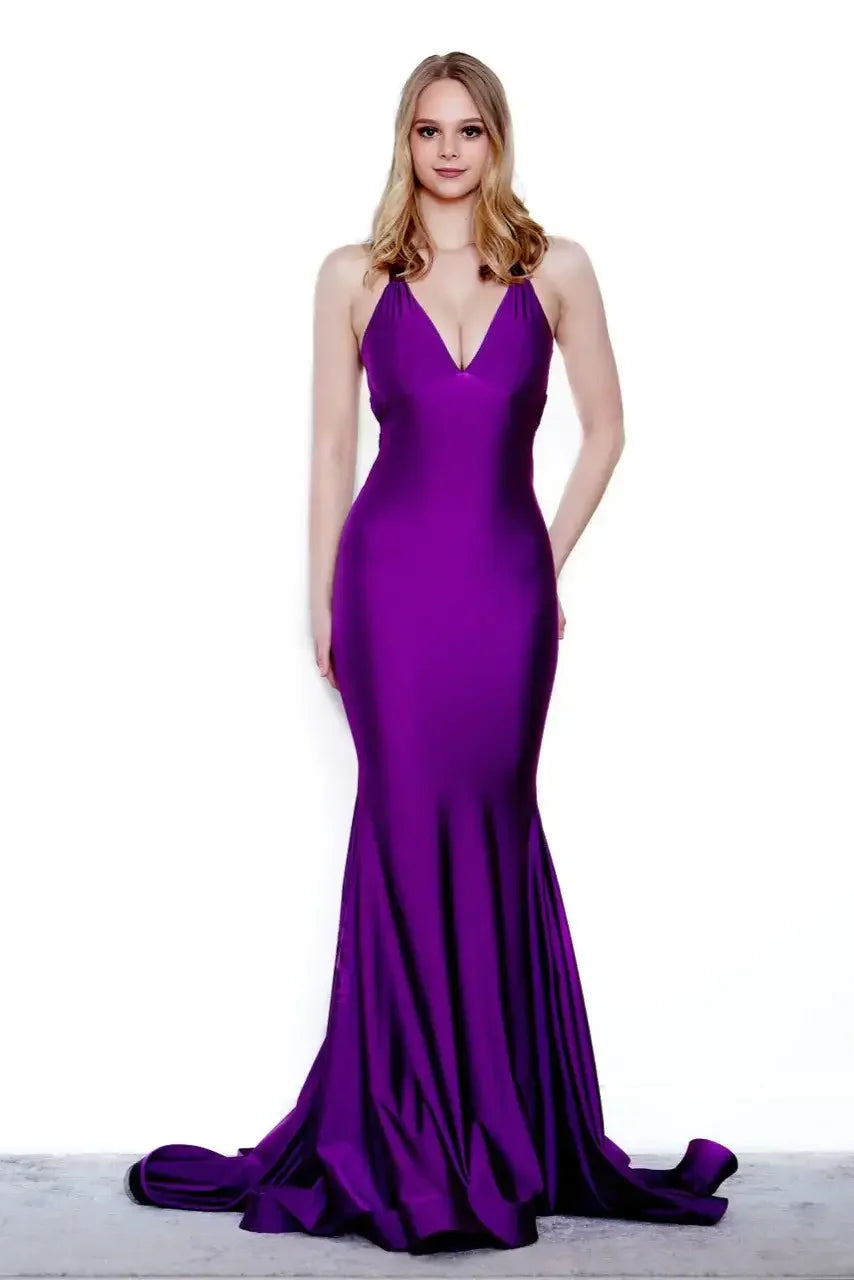A woman stands confidently in a long, fitted purple dress with a halter neck and a slight train, against a plain white background, exuding elegance and poise.