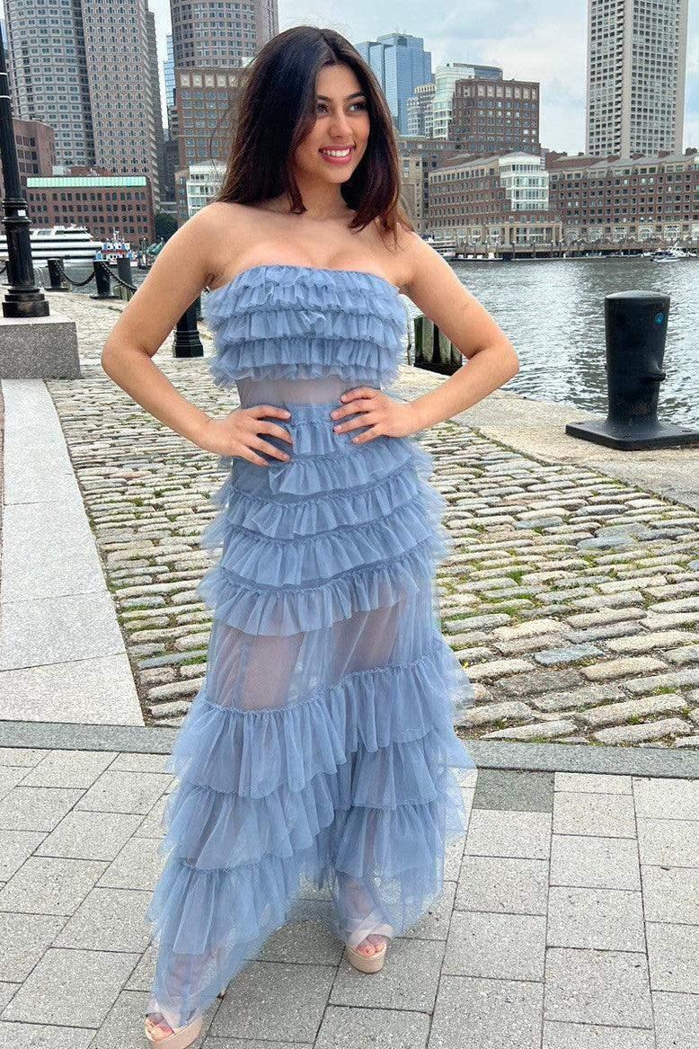 A woman stands by a waterfront in a layered, strapless light blue dress. She's smiling with her hands on her hips, surrounded by cobblestone paths and tall buildings in the background.