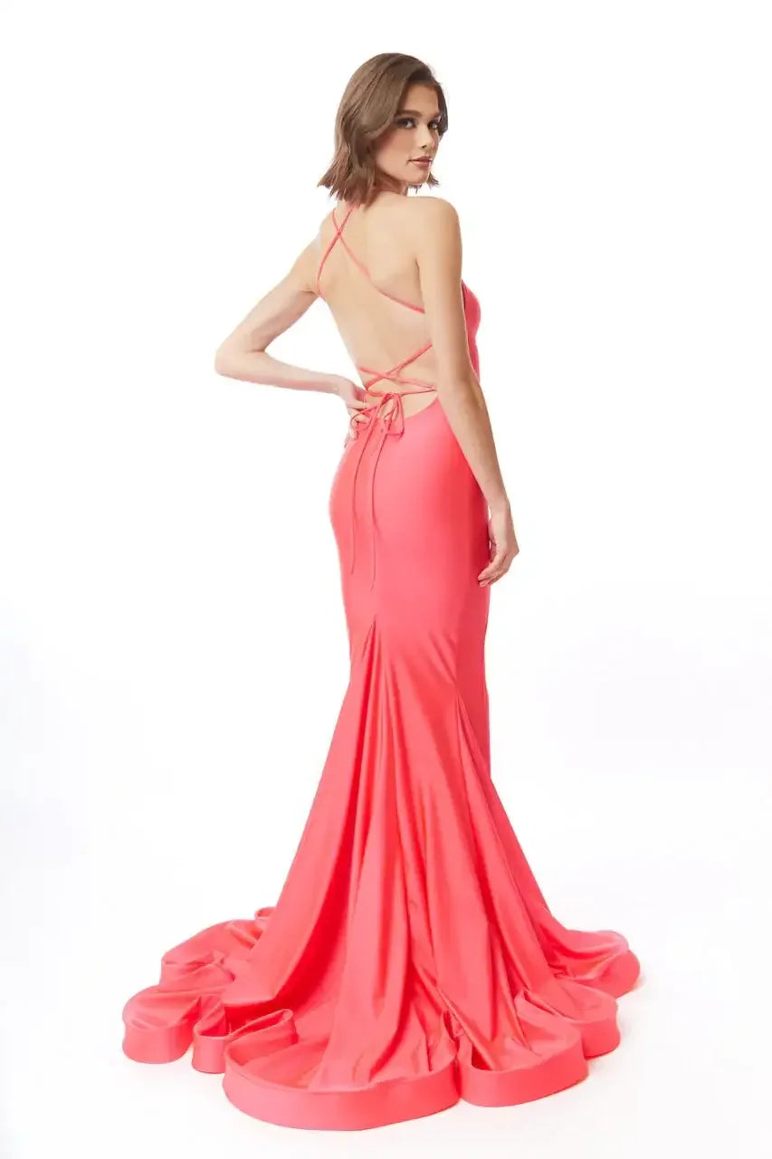 A woman stands elegantly in a fitted coral gown, showcasing an open back and flowing train. She poses gracefully against a plain white background, emphasizing the dress's vibrant color and design.