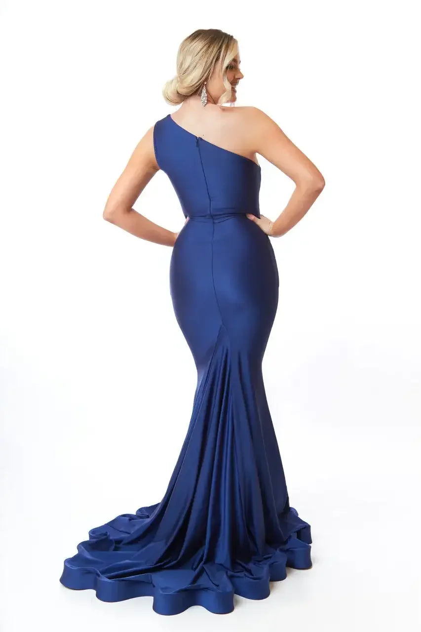 A woman poses with her back turned, wearing a fitted, one-shoulder navy gown that flares out into a dramatic ruffled train. The background is plain white.