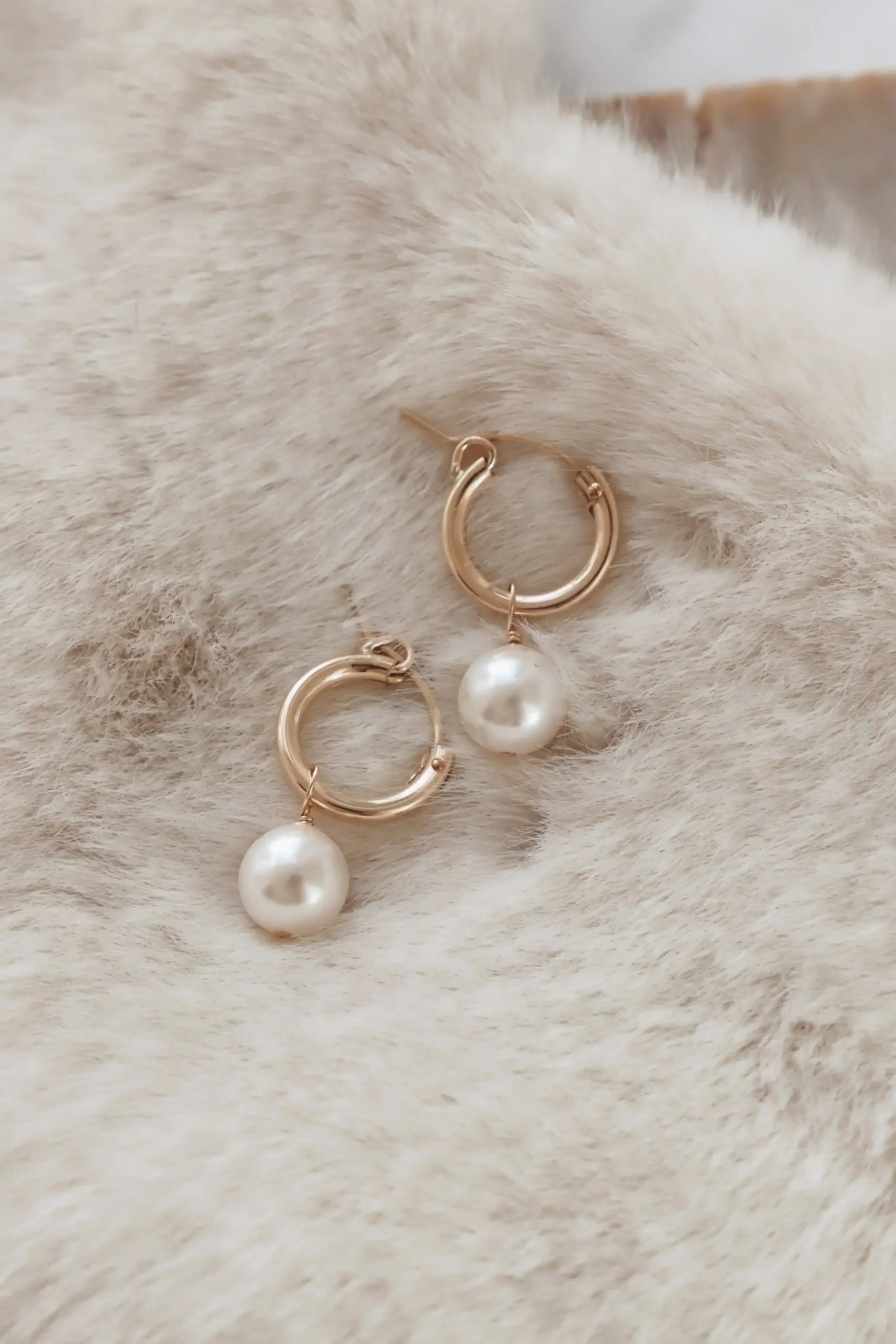 Gold hoop earrings with attached white pearls rest on a soft, textured cream-colored surface, creating an elegant display that highlights the jewelry's design and shine.