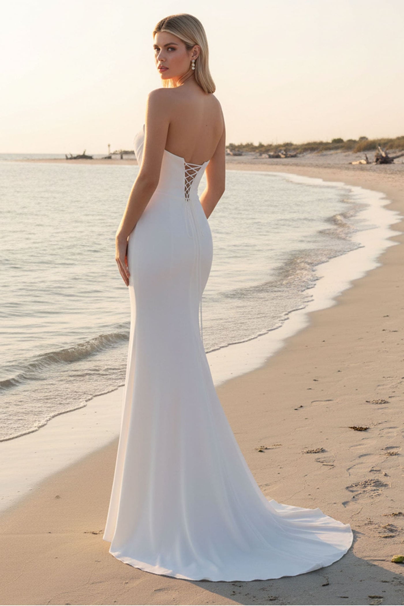 A woman stands on a sandy beach at sunset wearing a fitted white mermaid-style wedding gown with an open back and lace-up detail. The dress has a slight train that pools on the sand beside the shoreline.