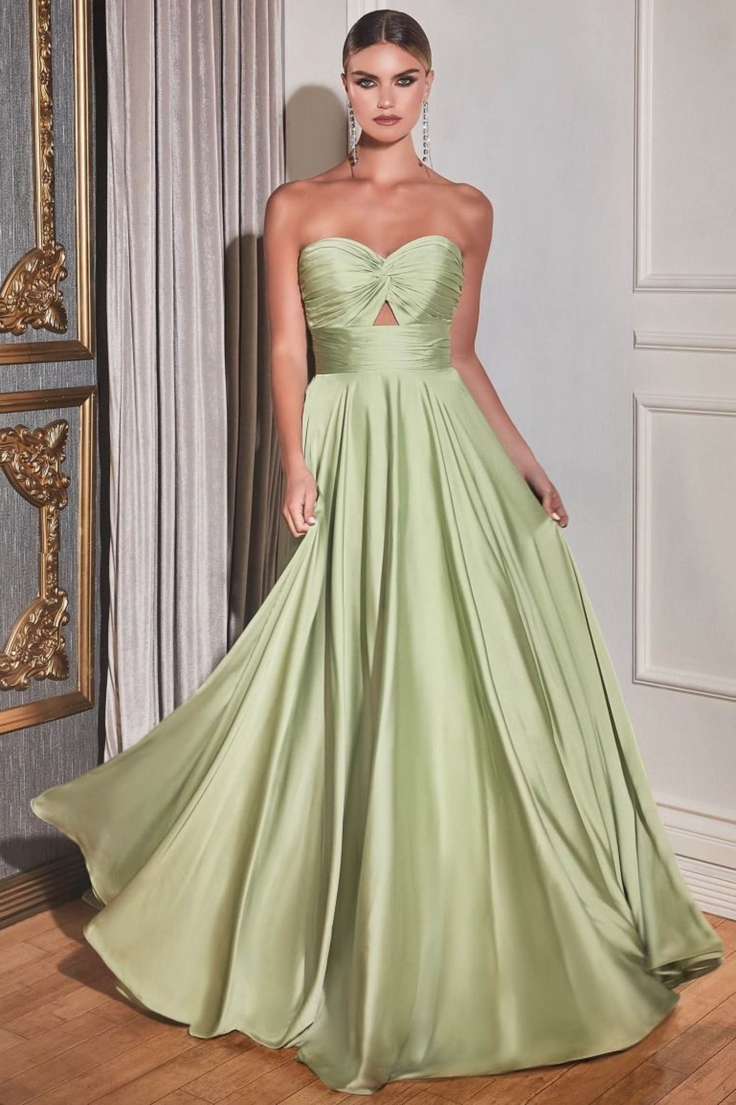 A woman models a strapless light sage green ball gown with a sweetheart neckline, a twisted bodice detail, and a full flowing skirt in an elegant indoor setting.
