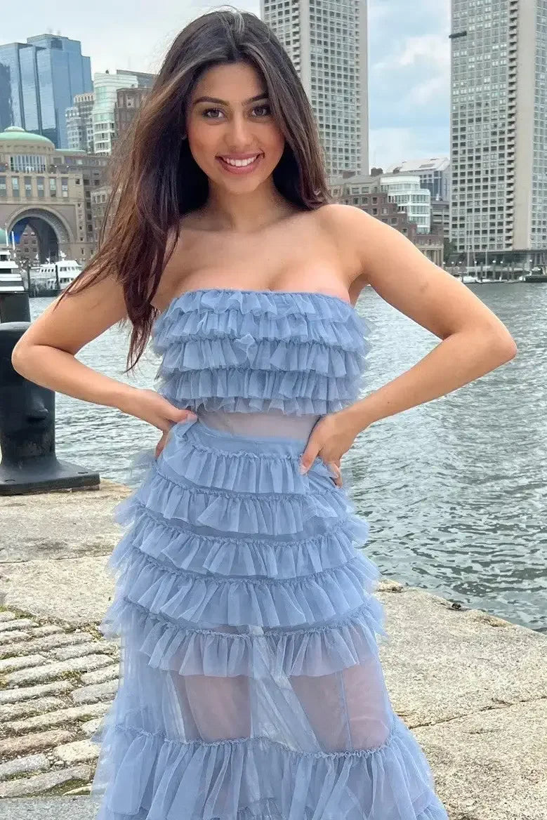 A woman stands on a cobblestone waterfront, smiling and posing in a blue ruffled dress. Behind her are modern buildings and boats on the water, under a cloudy sky.