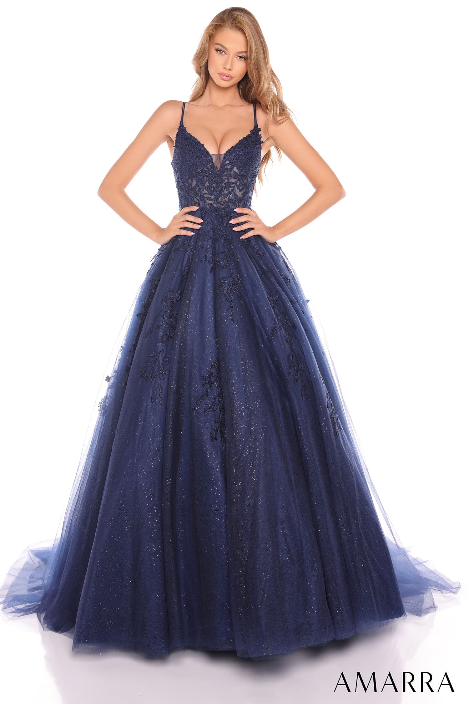 A woman modeling a navy blue ball gown with a lace bodice, thin spaghetti straps, and a voluminous tulle skirt against a white studio background. The brand name 'AMARRA' is printed in the bottom right corner.
