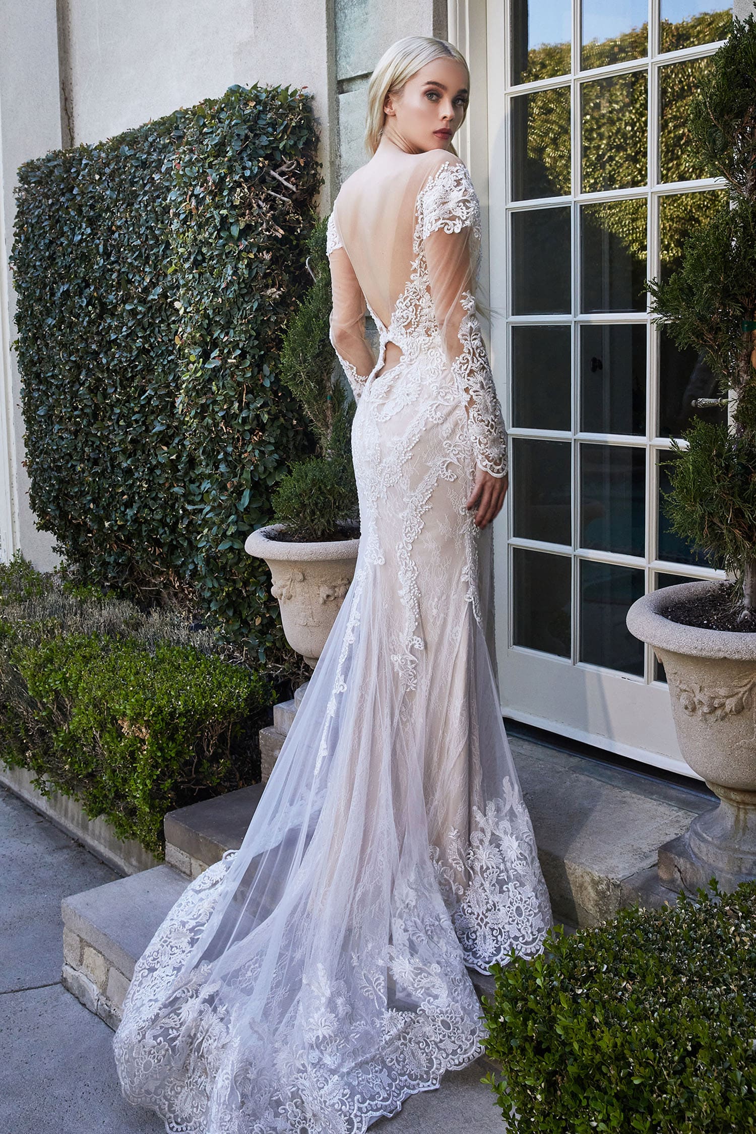 Woman in a white lace wedding dress standing outdoors with greenery around.