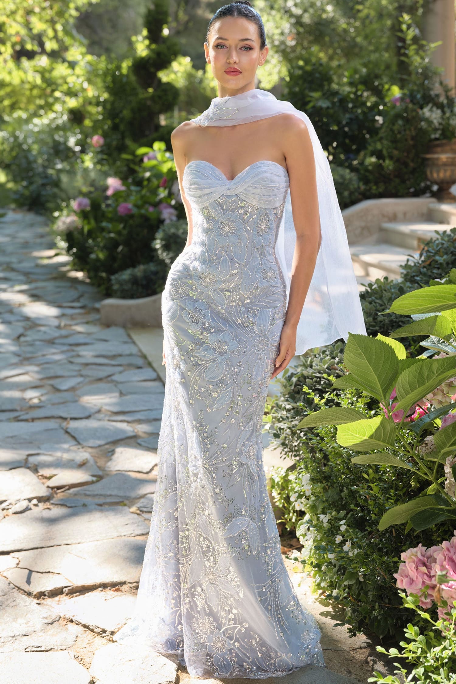 Woman in a white strapless gown with floral patterns standing outdoors on a stone path.