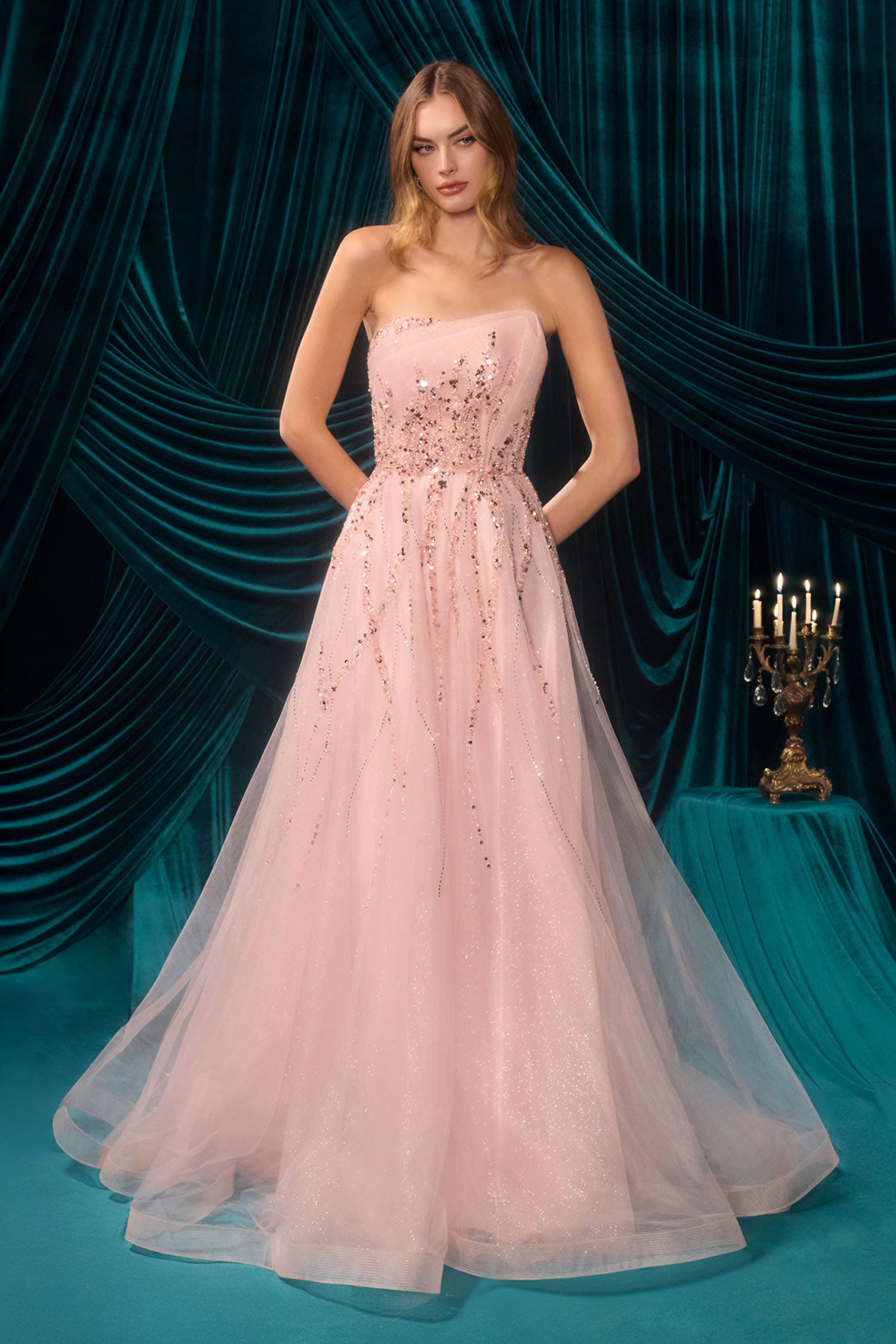 A woman models a strapless pale pink tulle gown with a heavily embellished beaded bodice, set against a dark teal velvet draped backdrop with a candelabra on a side table.
