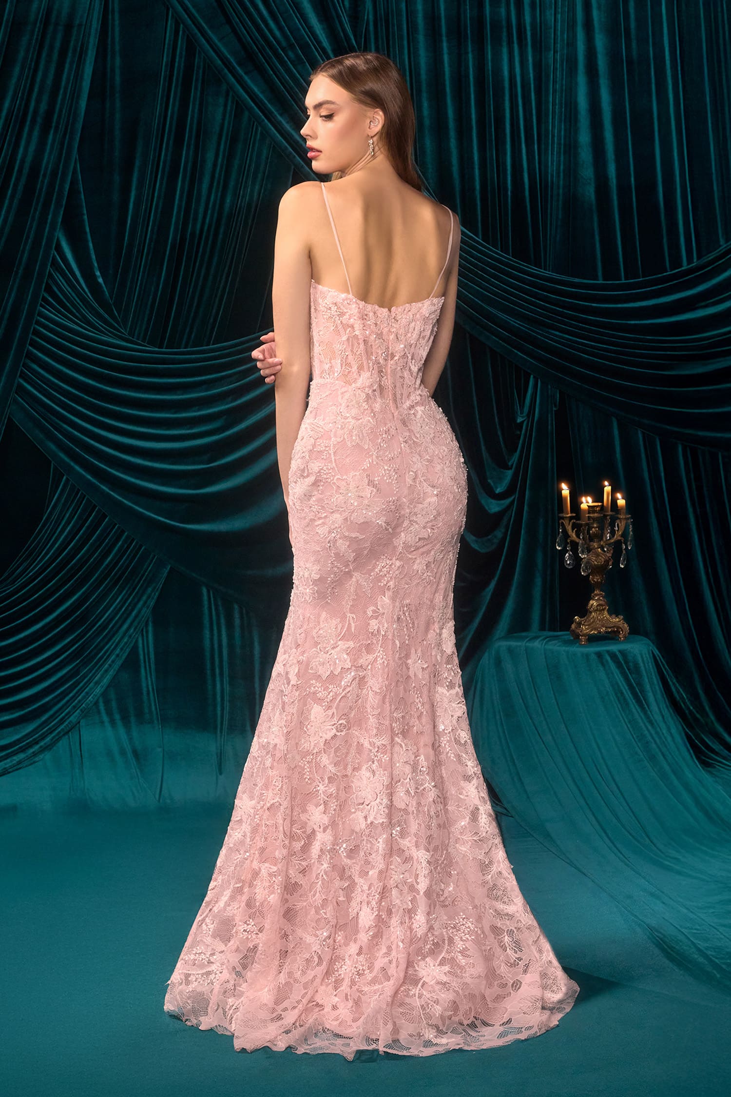 Woman in a pink lace evening gown standing against a dark teal curtain backdrop.