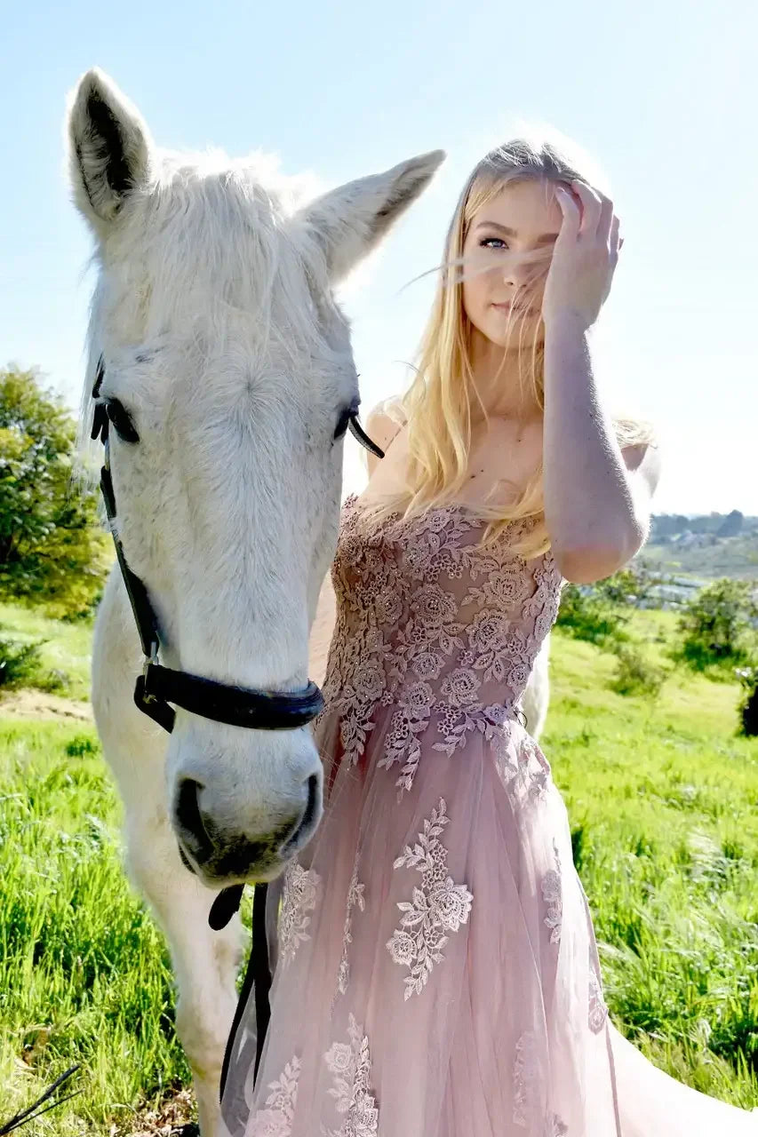 A young woman in a floral, light pink gown stands beside a white horse, gently touching her hair. They are in a lush, green outdoor setting under a clear blue sky.