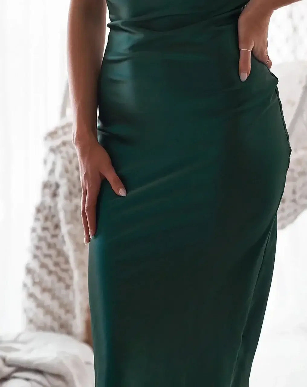 A sleek, green dress is the focal object, with a hand resting on the hip. The soft, bright environment suggests a cozy indoor setting, possibly near a window with drapes.