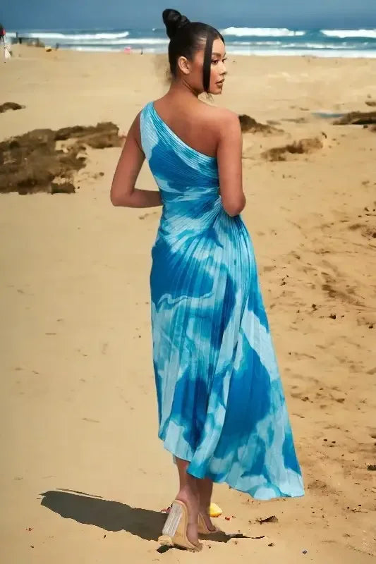 A woman in a flowing blue dress walks barefoot along a sandy beach. The ocean waves are visible in the background under a clear sky, creating a relaxed atmosphere.