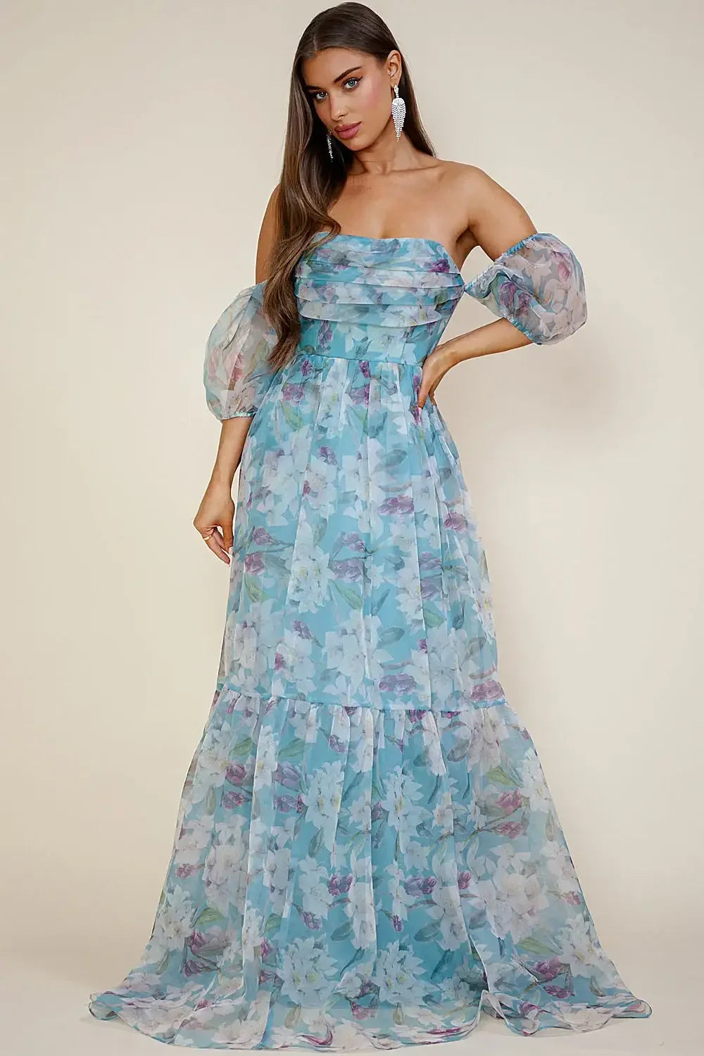 A stylish woman stands confidently wearing a flowing, floral-printed dress with off-the-shoulder sleeves. The light fabric cascades down, blending with a soft, neutral background. She poses elegantly, showcasing the dress.
