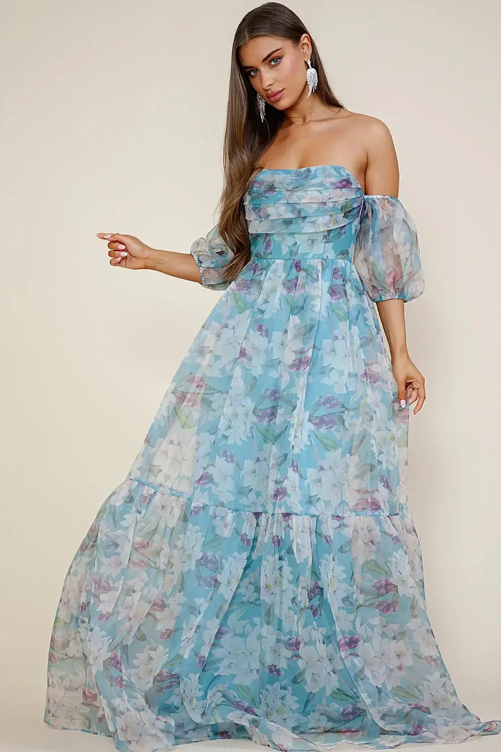 A woman wears a flowing, floral-patterned maxi dress with puffed sleeves, gesturing gracefully. The soft background complements the light fabric, enhancing the dress's delicate, romantic style.