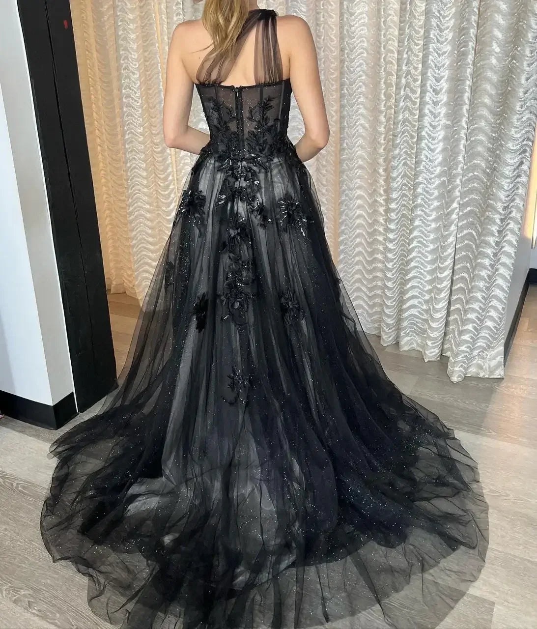 A woman in a black, floor-length evening gown adorned with floral embellishments poses, showcasing the dressâ€™s intricate details against a backdrop of sheer, draped curtains and wooden flooring.
