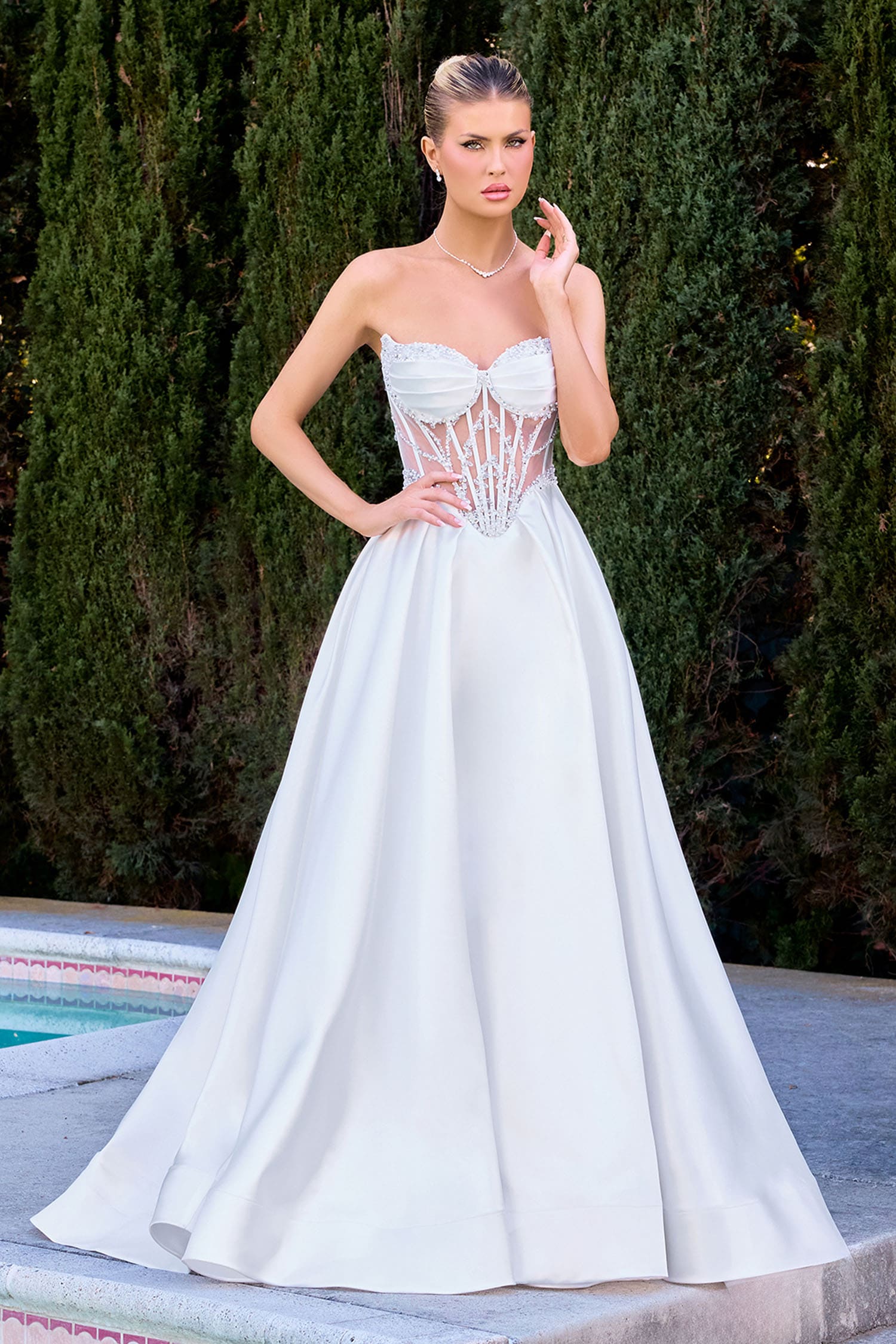 Woman in a white strapless gown standing by a pool with greenery in the background