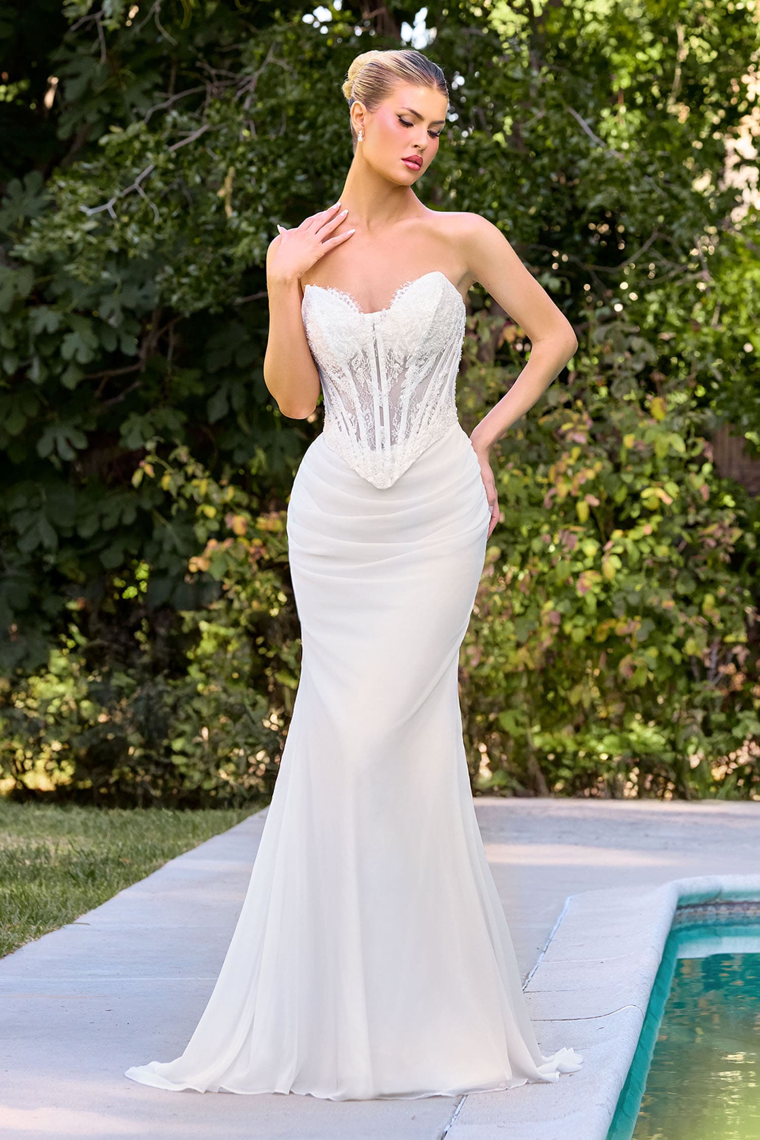 Woman in a white strapless gown standing outdoors near a pool with greenery in the background