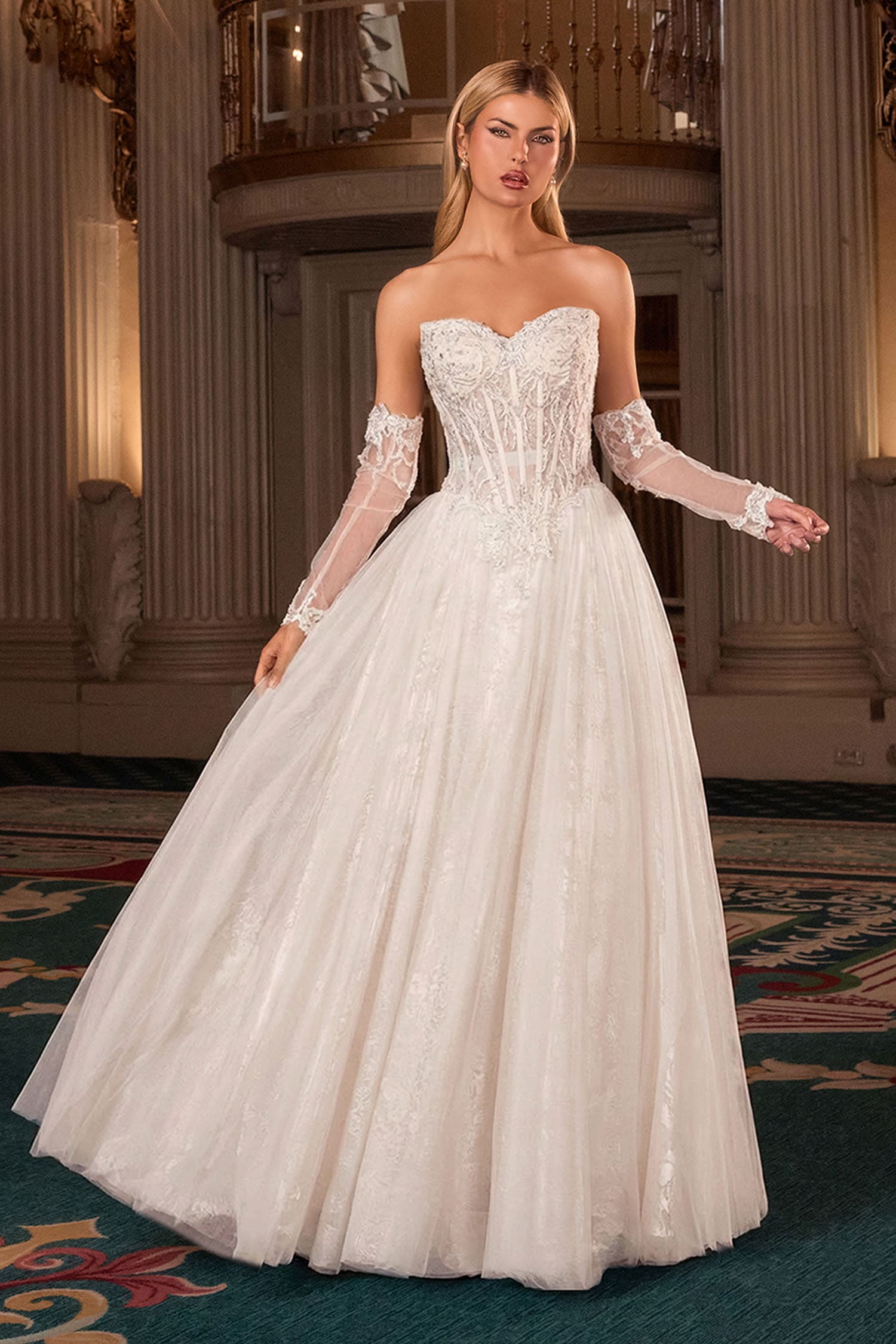 A woman modeling a strapless sweetheart ball gown wedding dress with long sheer lace sleeves, a fitted embellished bodice, and a voluminous tulle skirt in a grand indoor setting with classical columns.