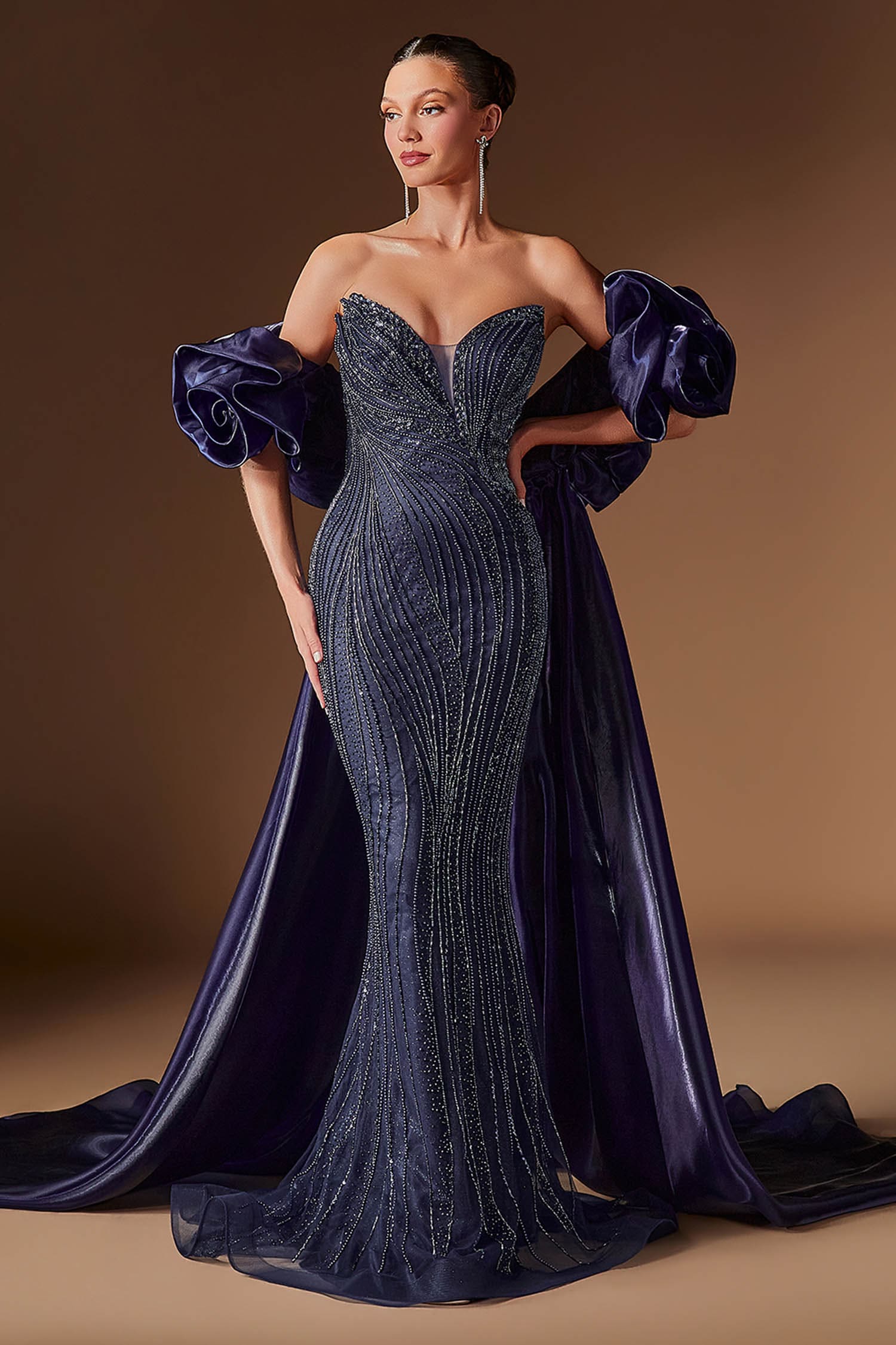A woman models a strapless evening gown with a sweetheart neckline, fitted beaded mermaid silhouette, dramatic puffed sleeves, and a long satin train against a warm brown studio background.