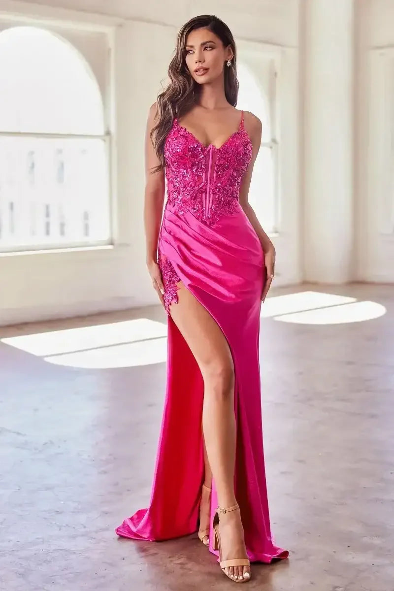A woman is posing elegantly in a vibrant pink dress with a thigh-high slit and detailed embellishments. She stands in a bright, spacious room with large windows, showcasing her stylish sandals.