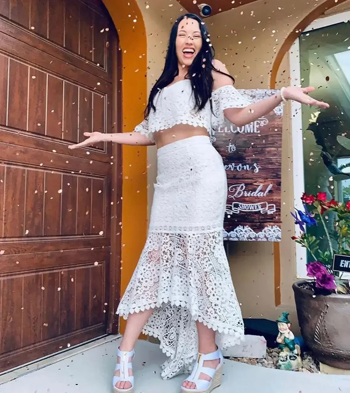A woman wearing a white lace outfit smiles joyfully with arms outstretched, surrounded by confetti. She stands in front of a door, with colorful flowers and decorations visible nearby.