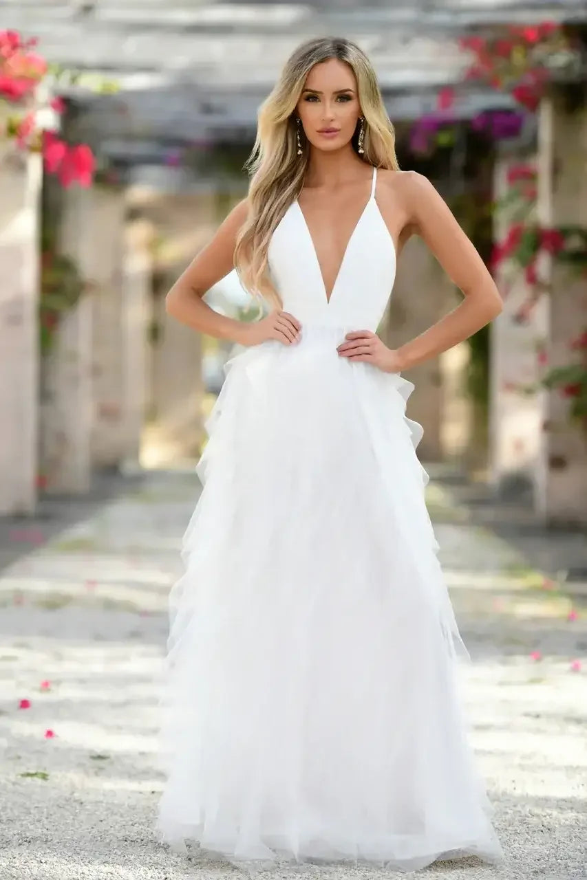 A woman stands confidently in a flowing white gown with a deep neckline. Soft pink and green floral decorations frame a picturesque outdoor setting, enhancing the serene atmosphere.