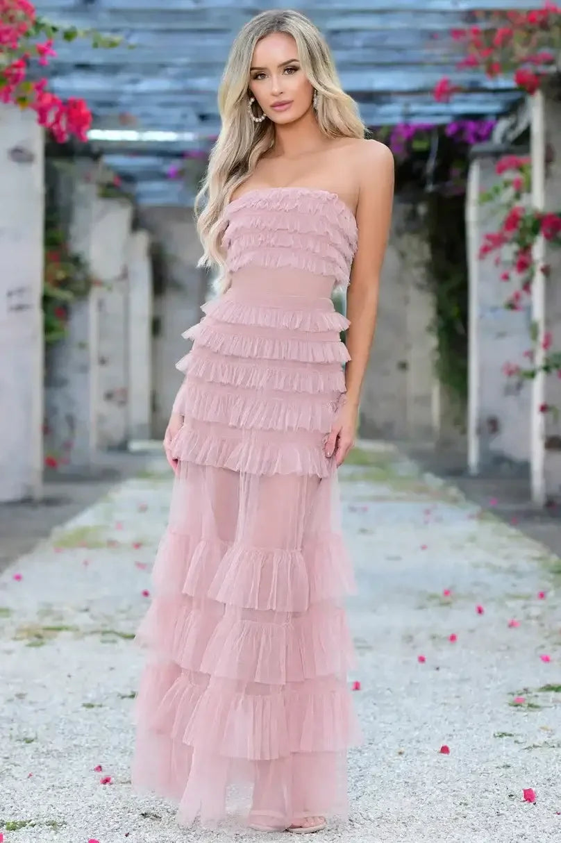 A woman in a layered, ruffled, light pink gown stands on a gravel path surrounded by blooming flowers. She poses confidently in a picturesque outdoor setting with stone structures.