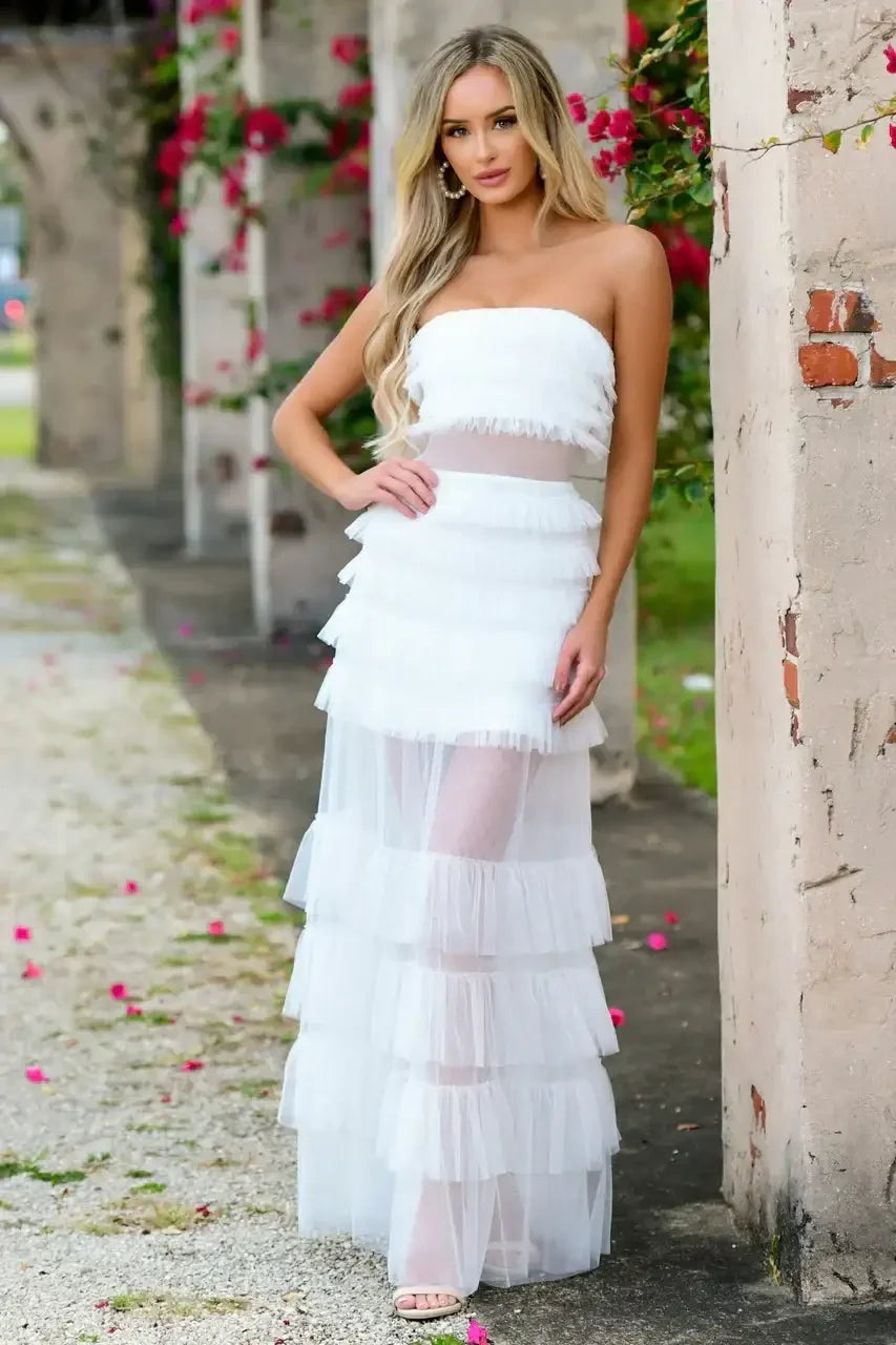 A woman stands confidently in a tiered white dress with a strapless top, surrounded by a scenic outdoor area featuring pink flowers and weathered stone columns.